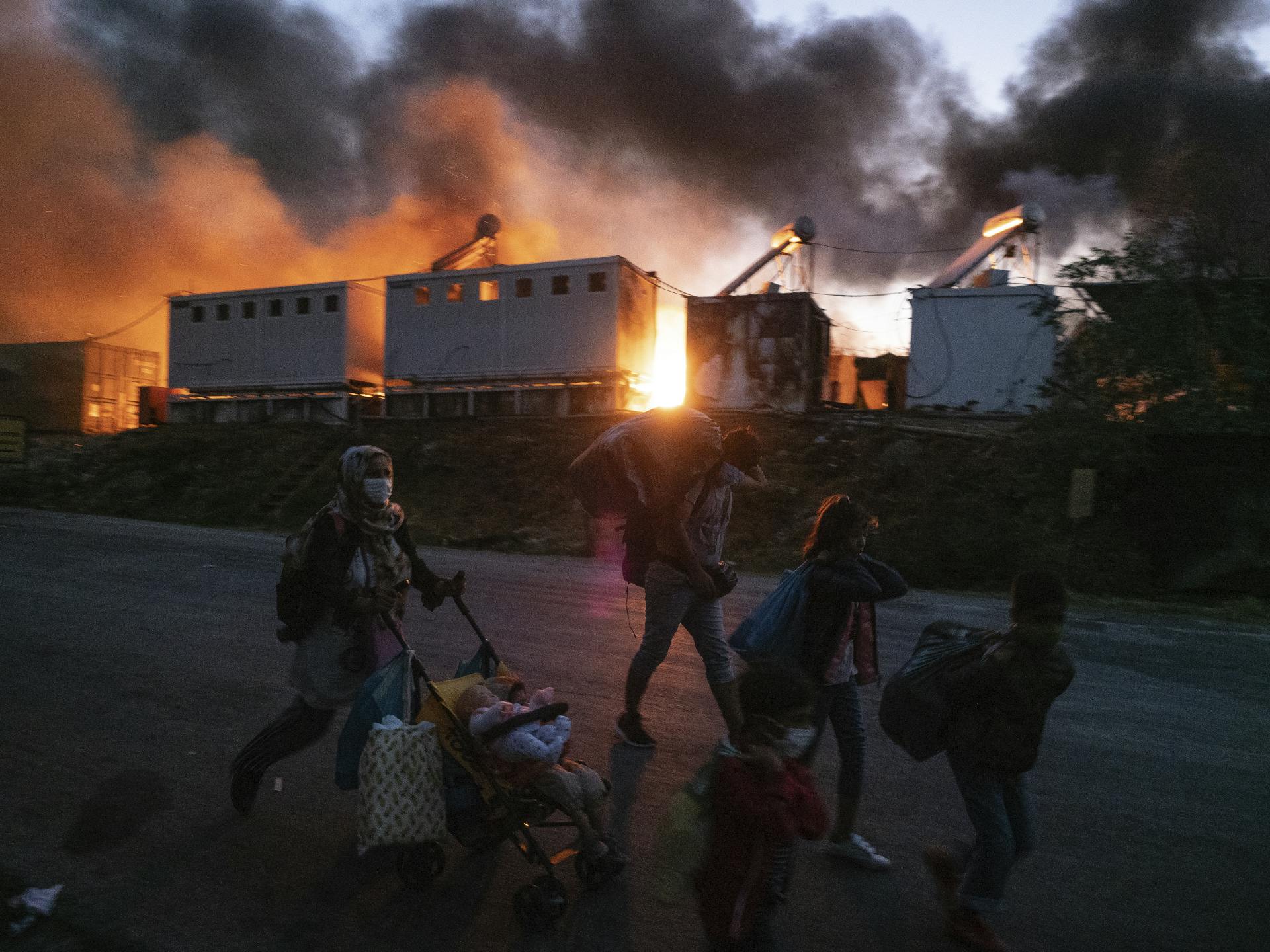 Refugees and asylum seekers walk with their possessions as a fire burns behind them in Lesbos, Greece. Photography by Enri Canaj.