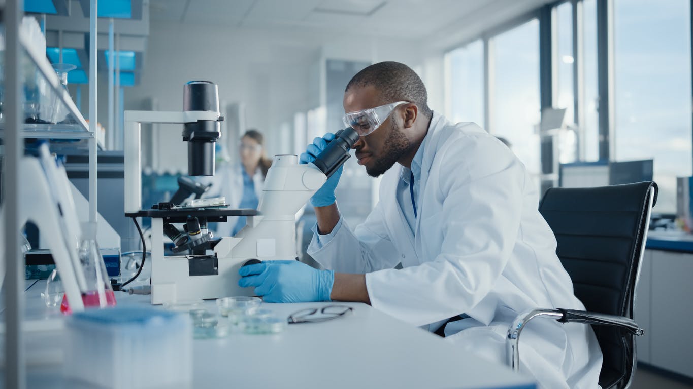A Sanofi researcher in a white robe looking through a microscope 