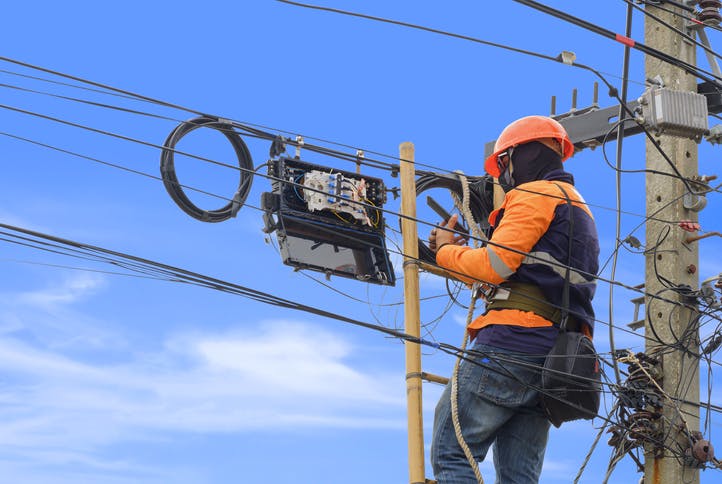 Technician using his phone to take a picture of an equipment, while standing on a ladder next to an electricity post