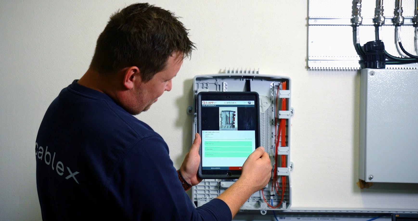 Cablex technician using a blue sweatshirt taking a photo of a fiber optic equipment with a tablet 