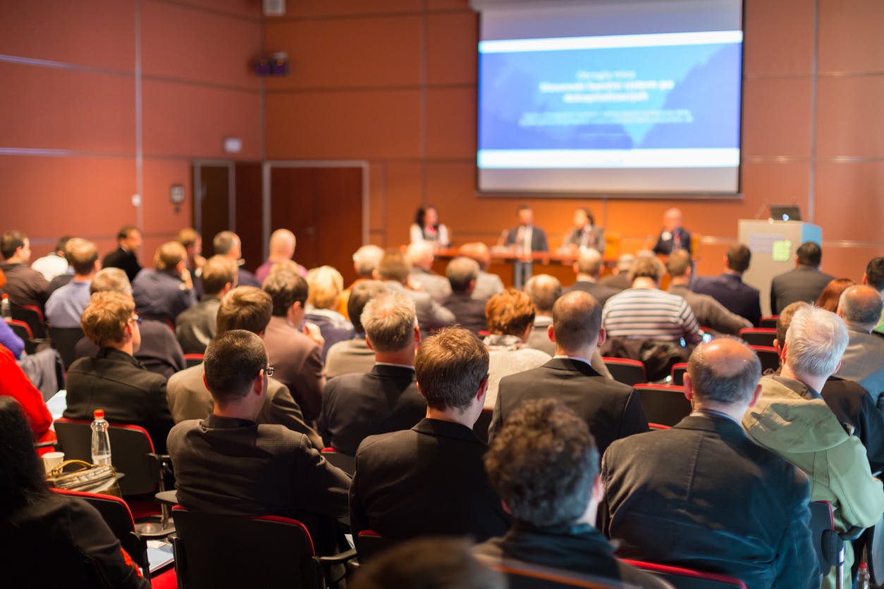 A group of adult workers sitting on red chairs attending to a Computer Vision Conference with four speakers