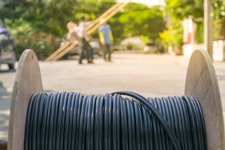 Close-up of a cable spool with workers carrying ladders in the background on a sunny day.