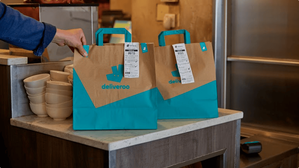 Two Deliveroo paper bags with printed receipts on a restaurant counter ready for collection.