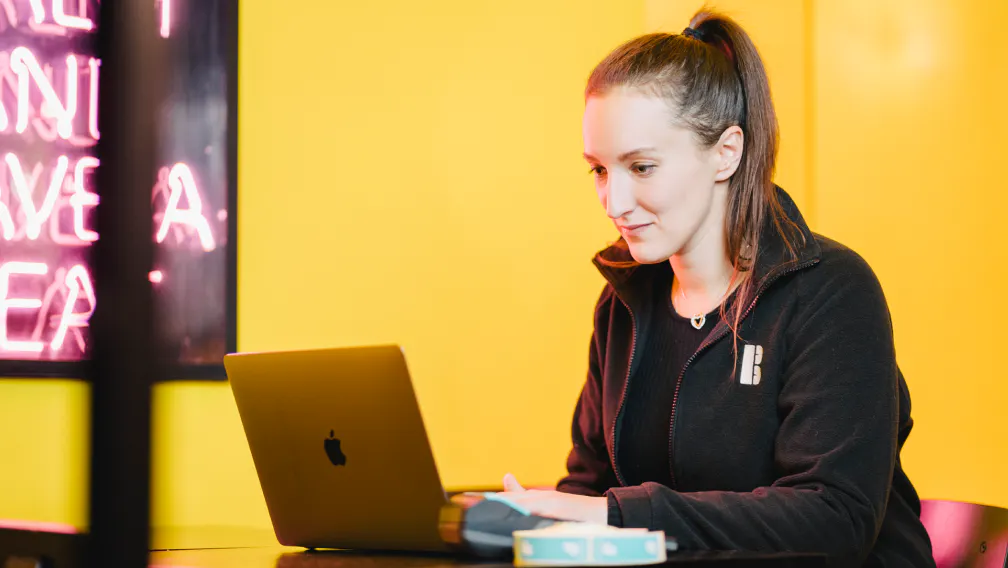 A woman with a high ponytail sits at a table in a brightly lit yellow room, focused on her laptop. She’s wearing a black zip-up jacket with a small white logo on the chest, and there’s a pink neon sign glowing in the background.