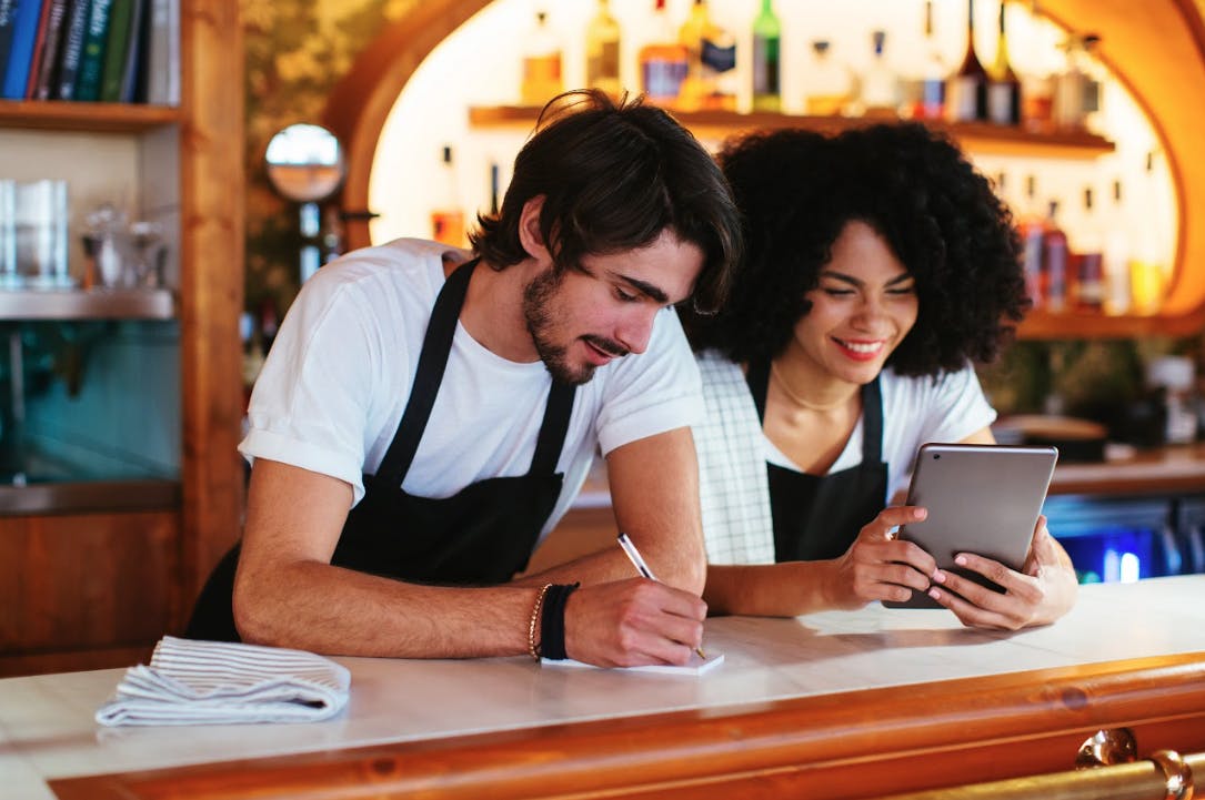 Two restaurant staff stand behind a counter, reviewing menu details on a tablet while one takes notes.