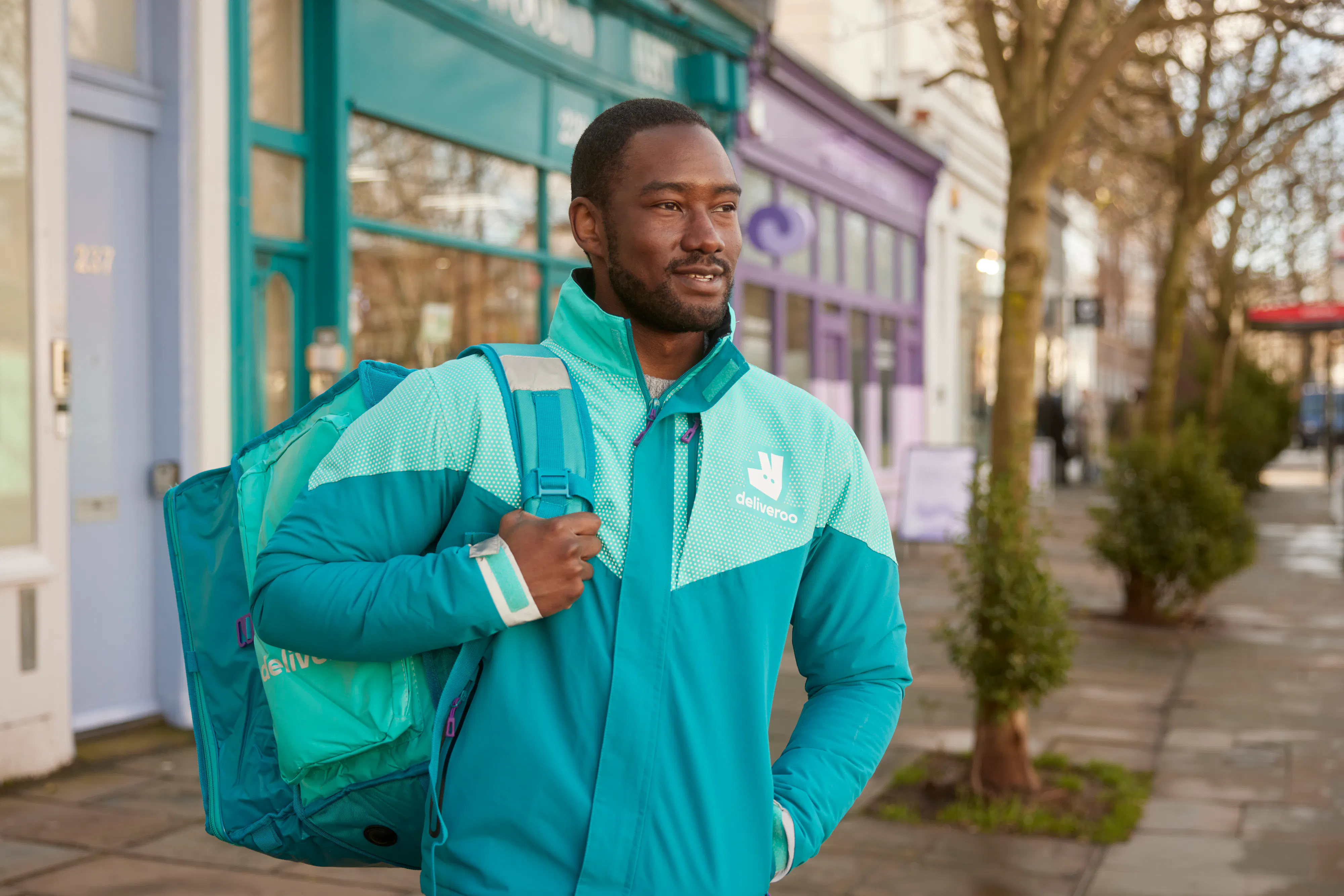 Man in Deliveroo coat with a Deliveroo delivery bag over his shoulder standing in front of shops