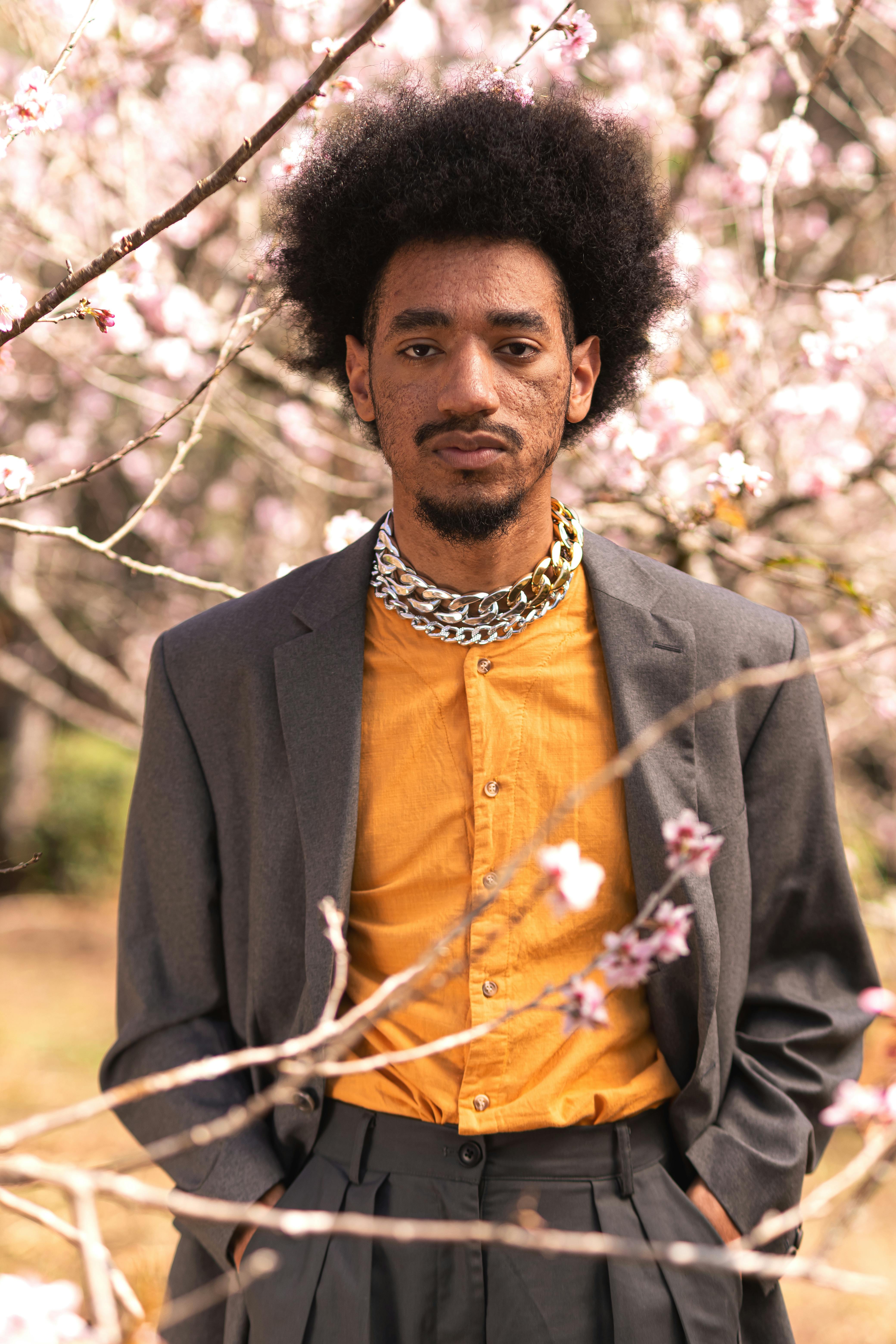 menino thito em pé a frente de uma árvore com flores rosas, vestido com uma camisa amarela e um terno cinza acompanhados de um colar de correntes grossas
