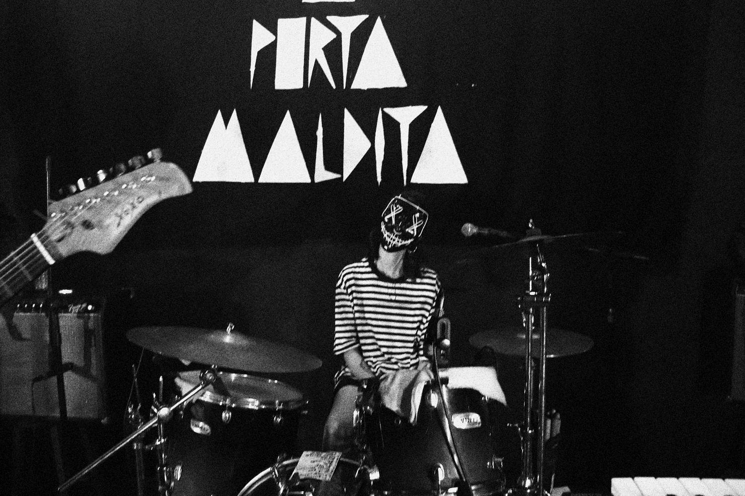 Baterista Nat sentada a frente de uma parede preta onde se lê o nome da casa "A Porta Maldita" em letras estilizadas geometricamente.