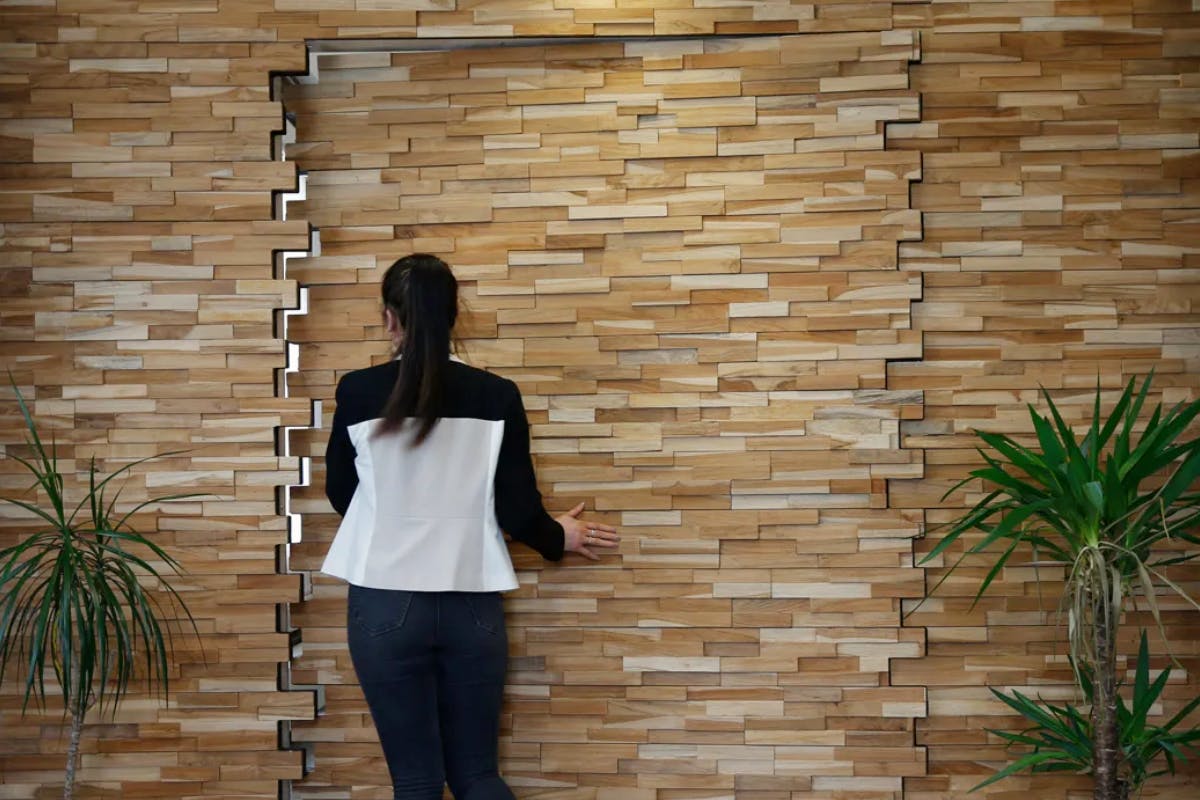 A woman entering through a bespoke secret pivot door by Deuren. The door and surrounding wall surfaces are finished in stacked timber blocks. The doors appear carved out of the blocks.