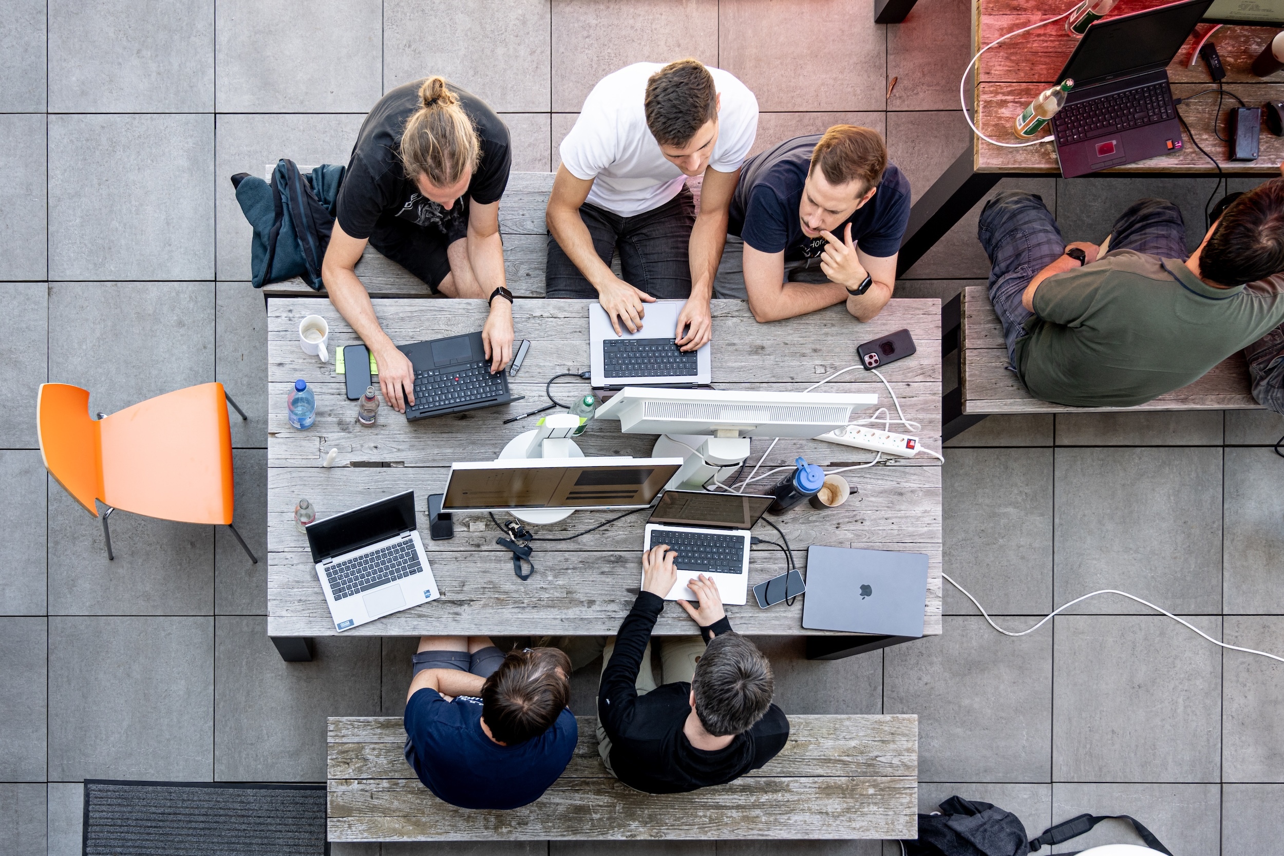 Luftaufnahme eines Entwicklerteams, das im Freien an einem Holztisch mit mehreren Laptops und Monitoren zusammenarbeitet.
