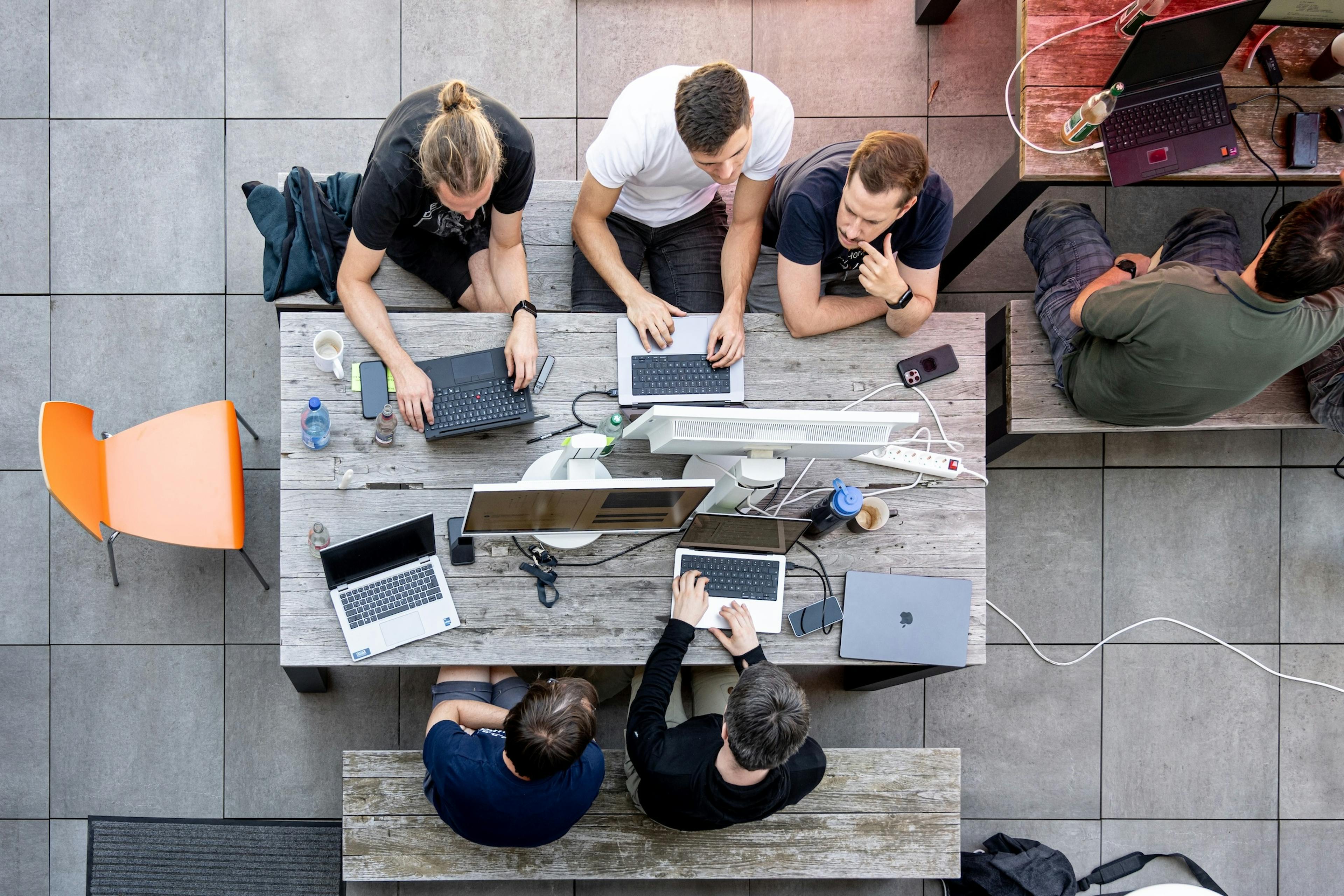 Luftaufnahme eines Entwicklerteams, das im Freien an einem Holztisch mit mehreren Laptops und Monitoren zusammenarbeitet.
