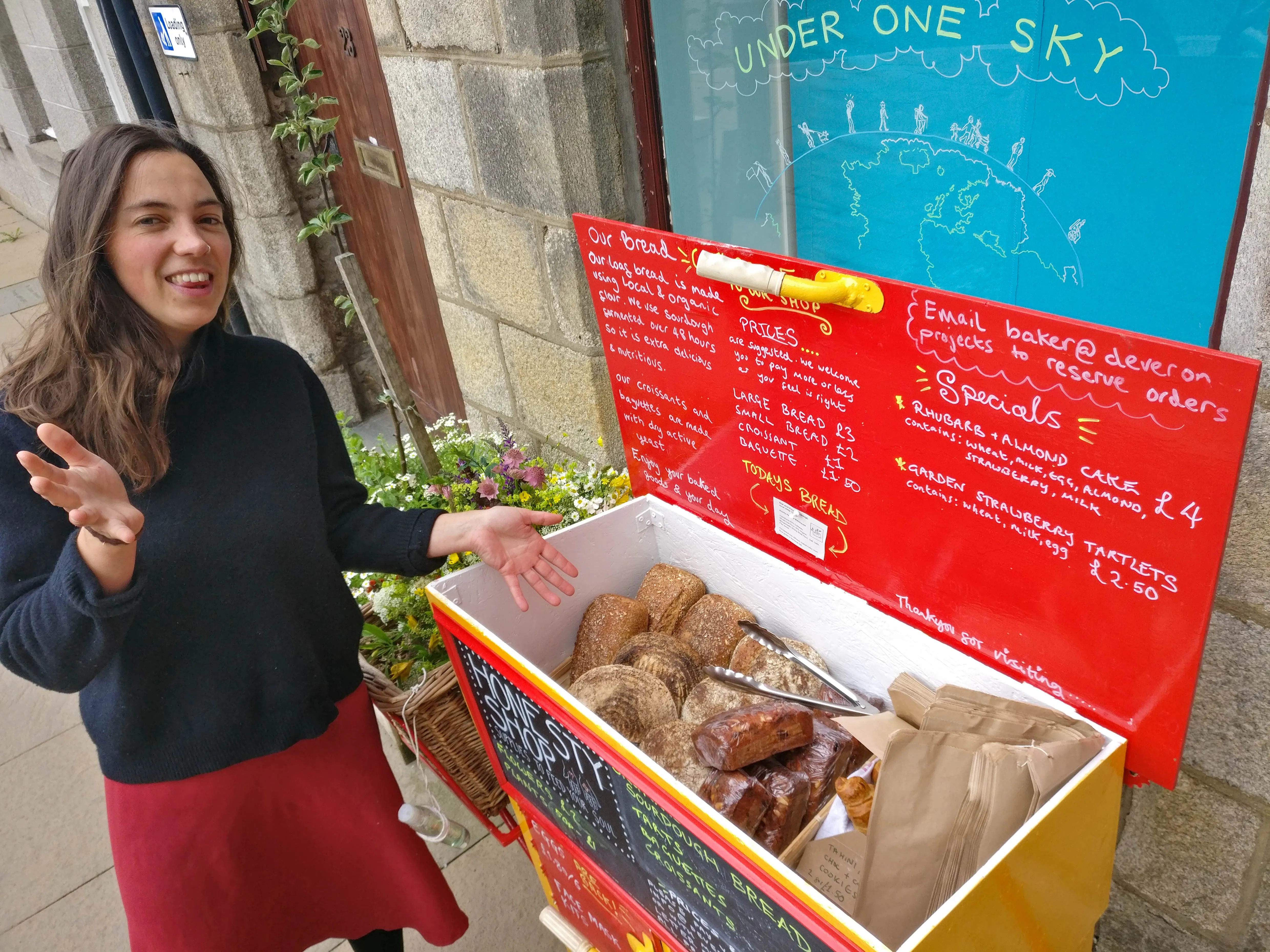 A light skinned woman with long brown har stands beside a wooden box painted red and yellow. They are located on  a street with stone buildings in the background. The box has a blackboard sign with a list of items written in chalk. 