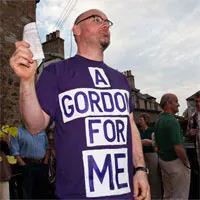 A light skinned man with balled head and dark framed glasses stands on a street wearing a purple T-shirt with a slogan that reads A GORDON FOR ME.