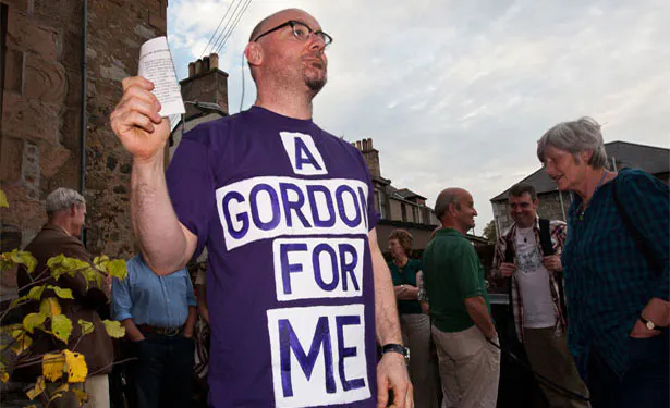 A light skinned man with balled head and dark framed glasses stands on a street wearing a purple T-shirt with a slogan that reads A GORDON FOR ME