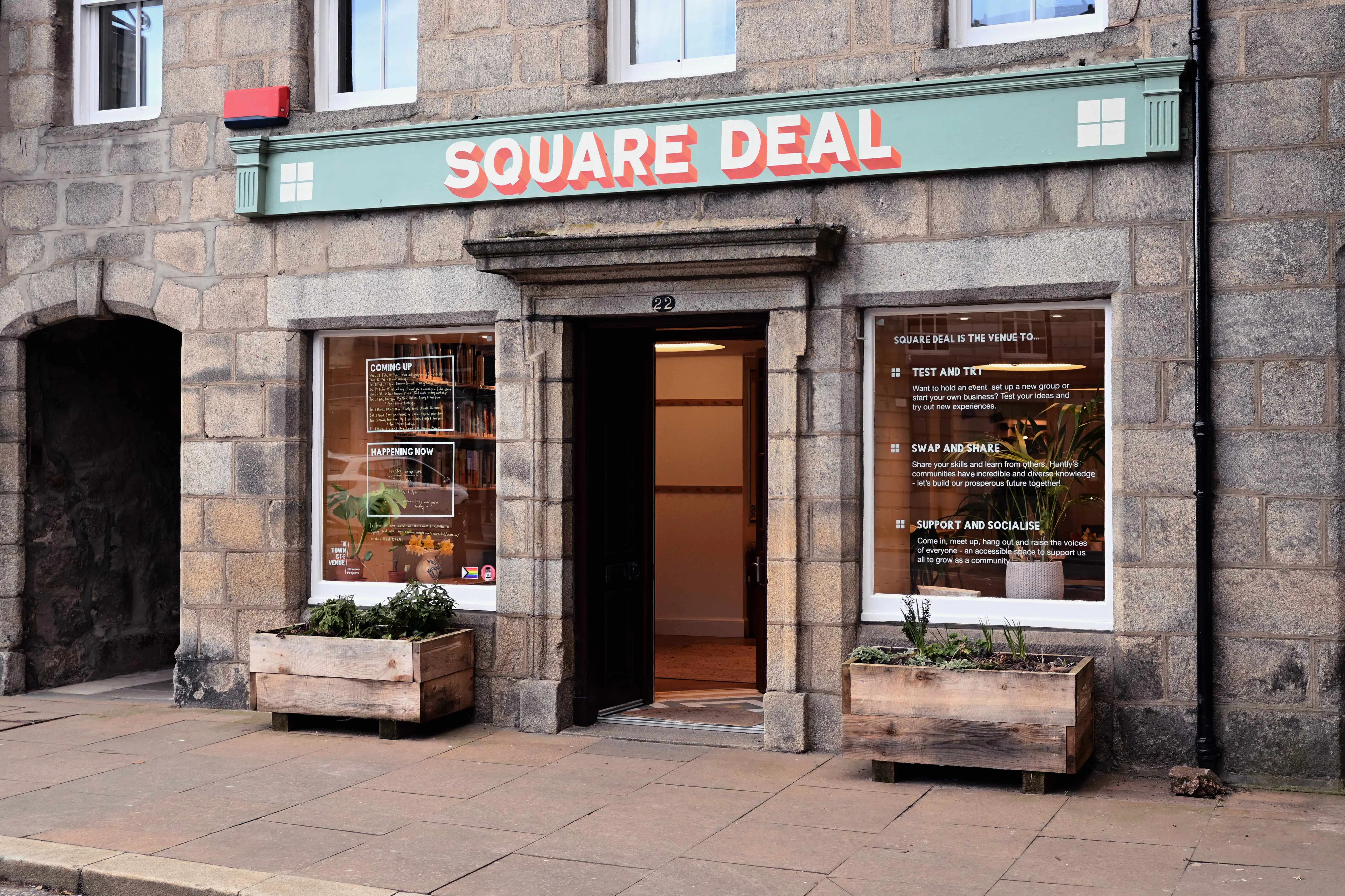 A photo showing the front of Square Deal community hub. It is a symmetrical stone building with two shop front windows either side of an open door. There is a large planter in front of each window.
