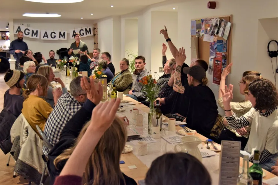 A photo of Deveron Projects' AGM in 2022. A long table is surrounded by people who are raising their hands.