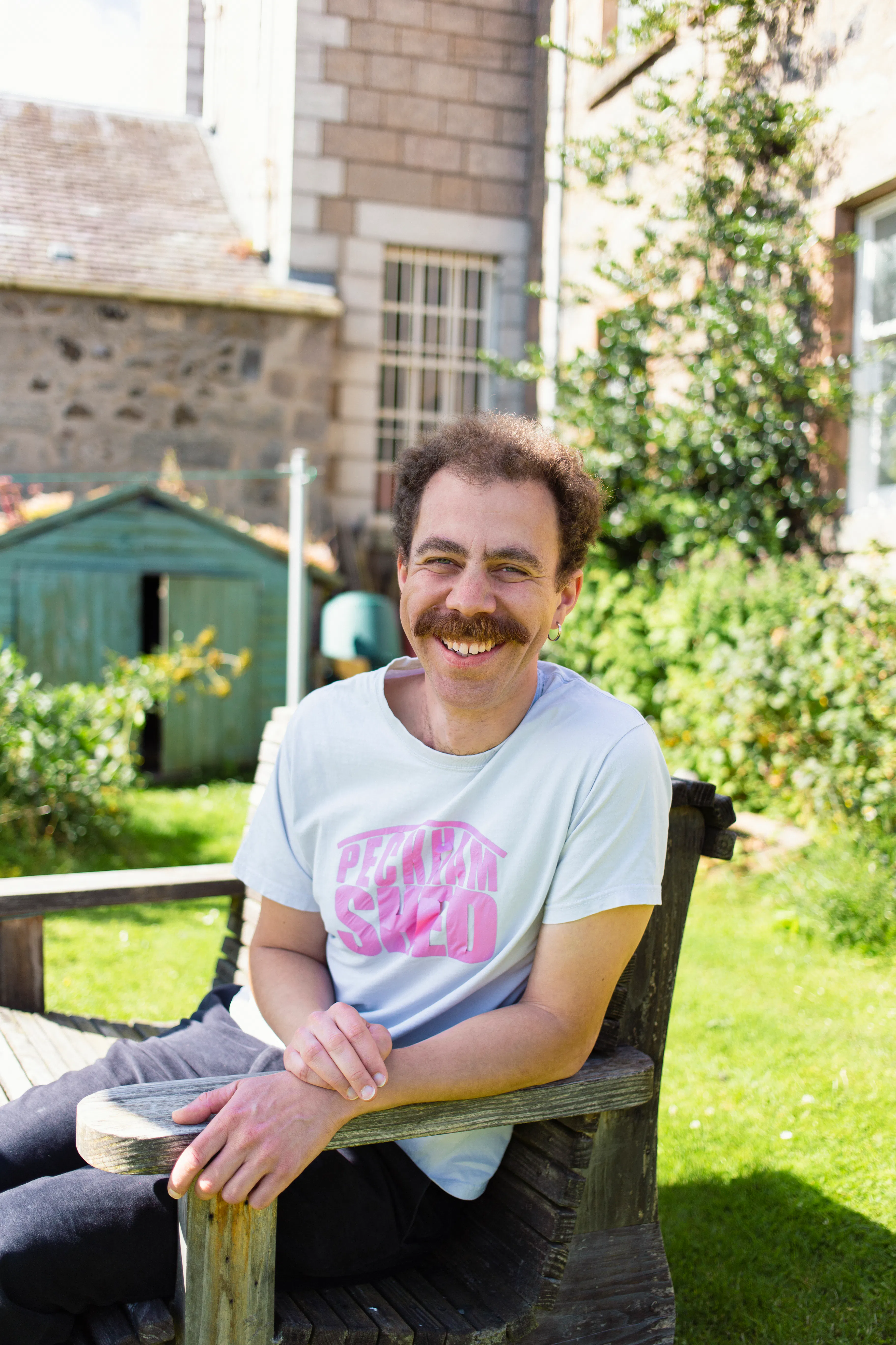 A man sits on a bench with a sunny garden behind him. He has light skin, dark curly hair and a moustache. He wears a white T-shirt and dark trousers.