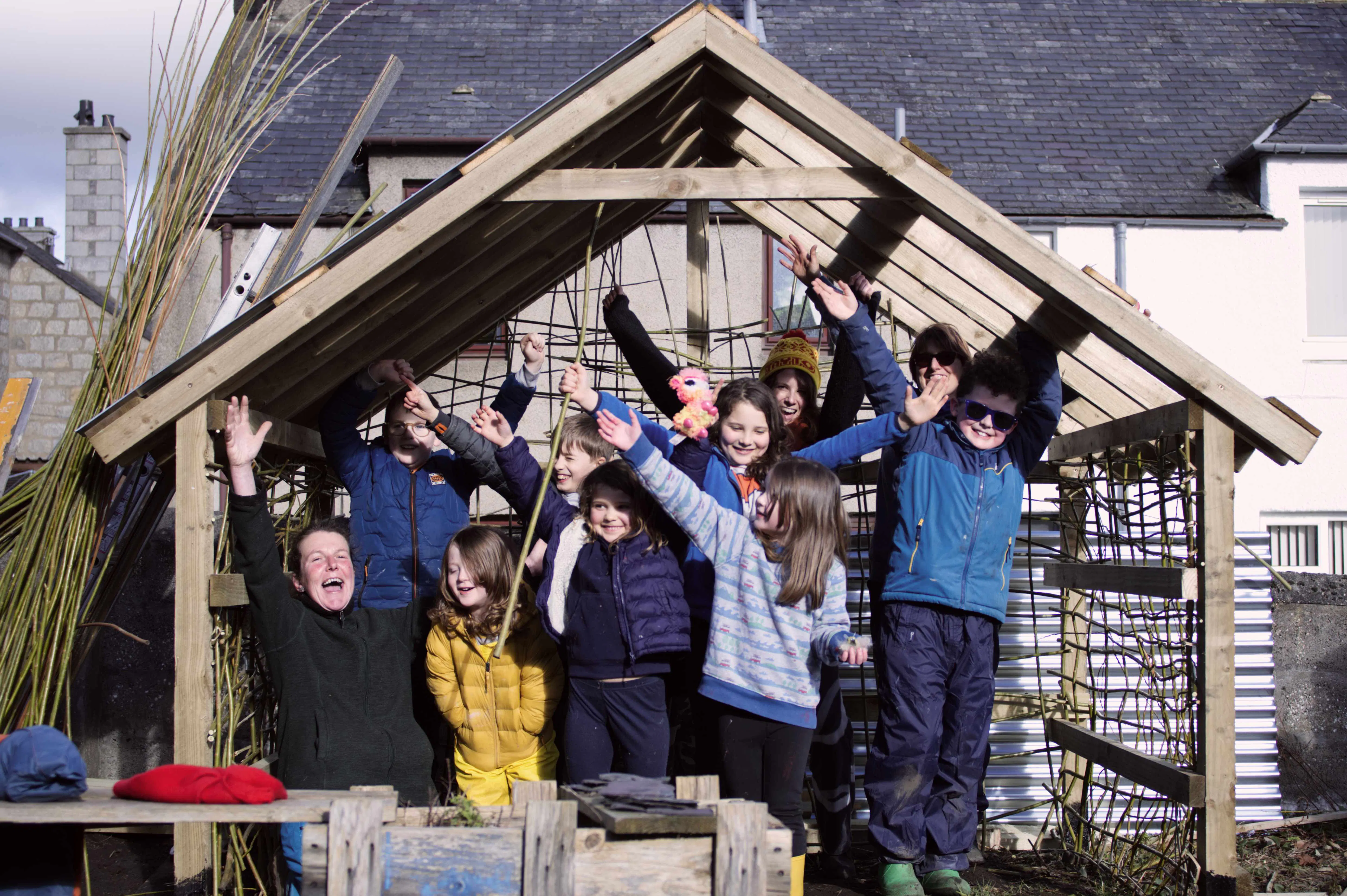 A group of children celebrate under a wooden shelter in the Brander Garden