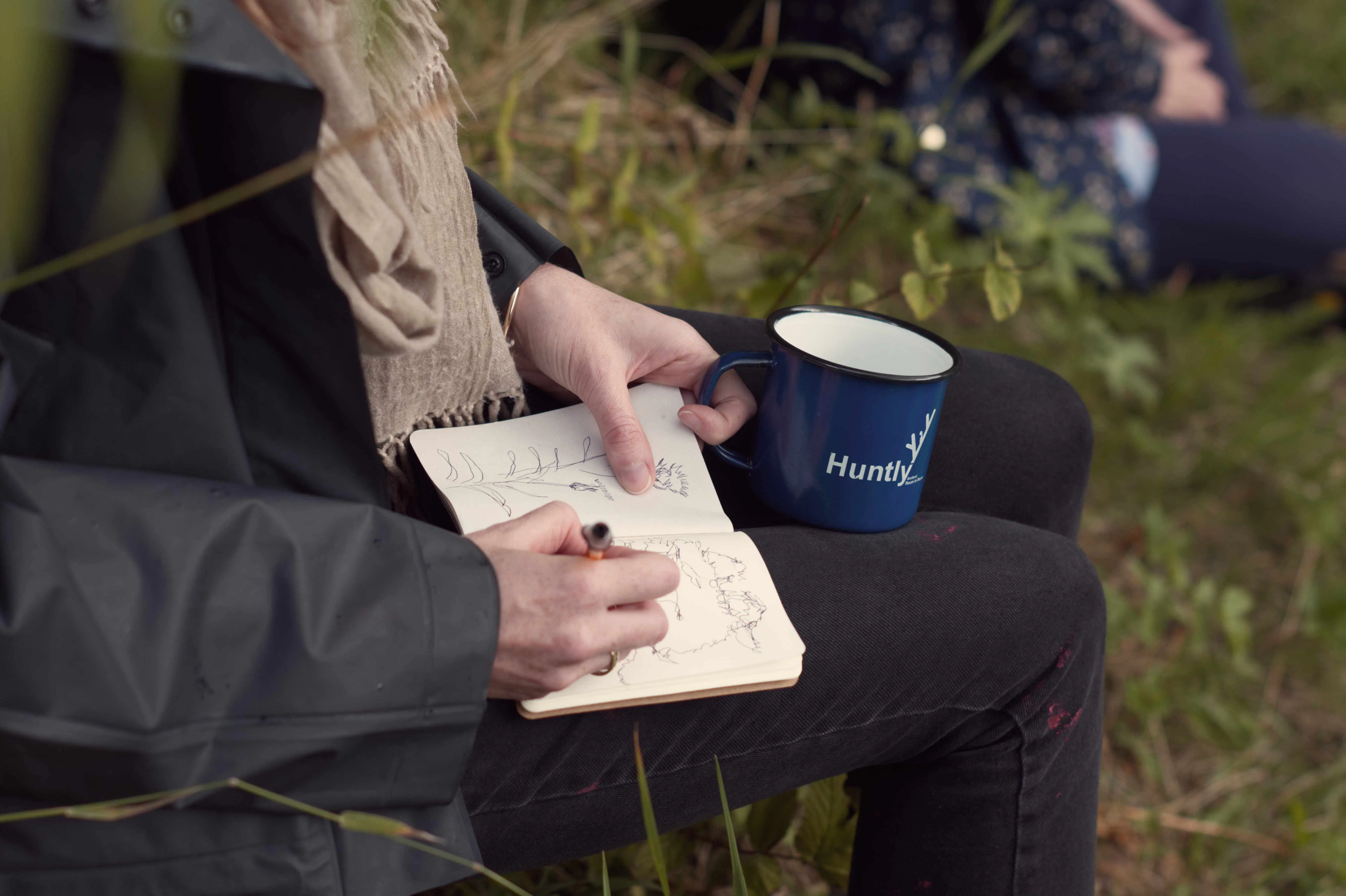 A person writes into a small notebook on their lap. They are holding a navy enamel cup with Huntly branding on it.