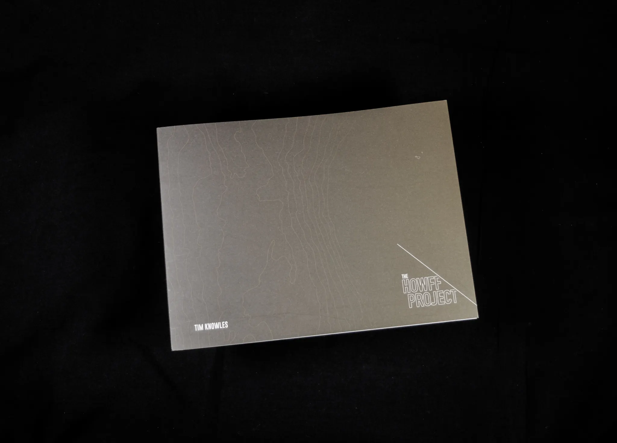The front cover of Tim Knowles' publication 'The Howff Project' on top of a black background. The cover is a dark grey, with faint contour lines. In the bottom left corner is Tim Knowles' name in white capitalised letters, and in the bottom right corner 'The Howff Project' is written underneath a diagonal line. 