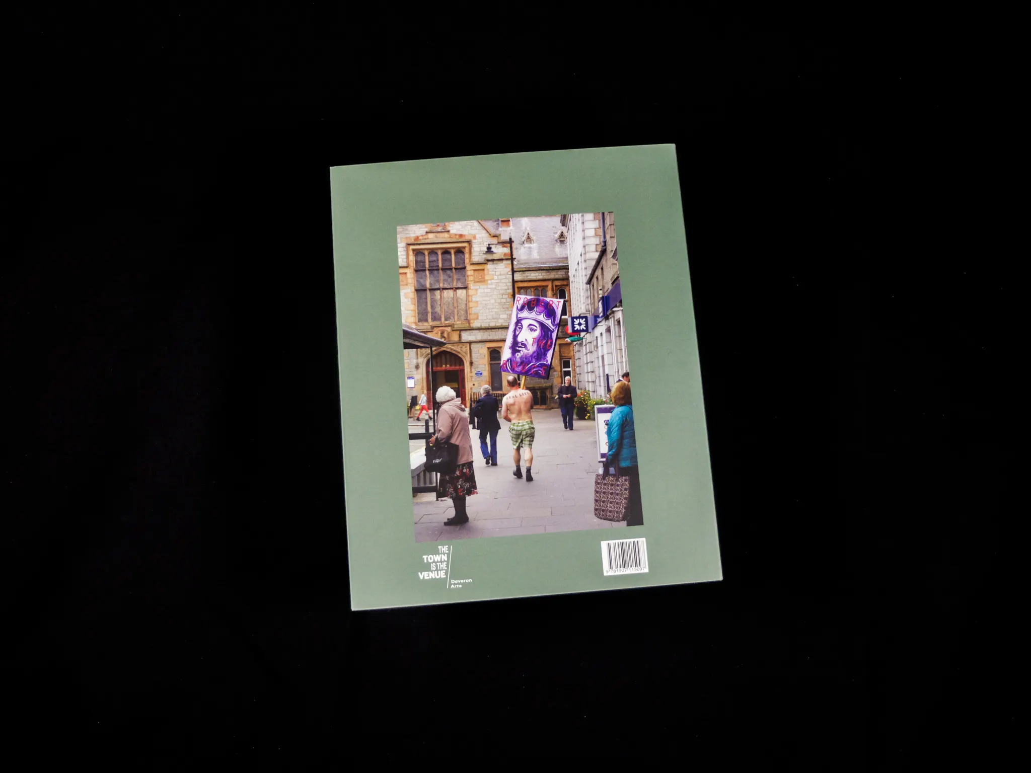The back cover of a book, featuring a photograph of a person walking through a busy Huntly street with a large painting held above their head