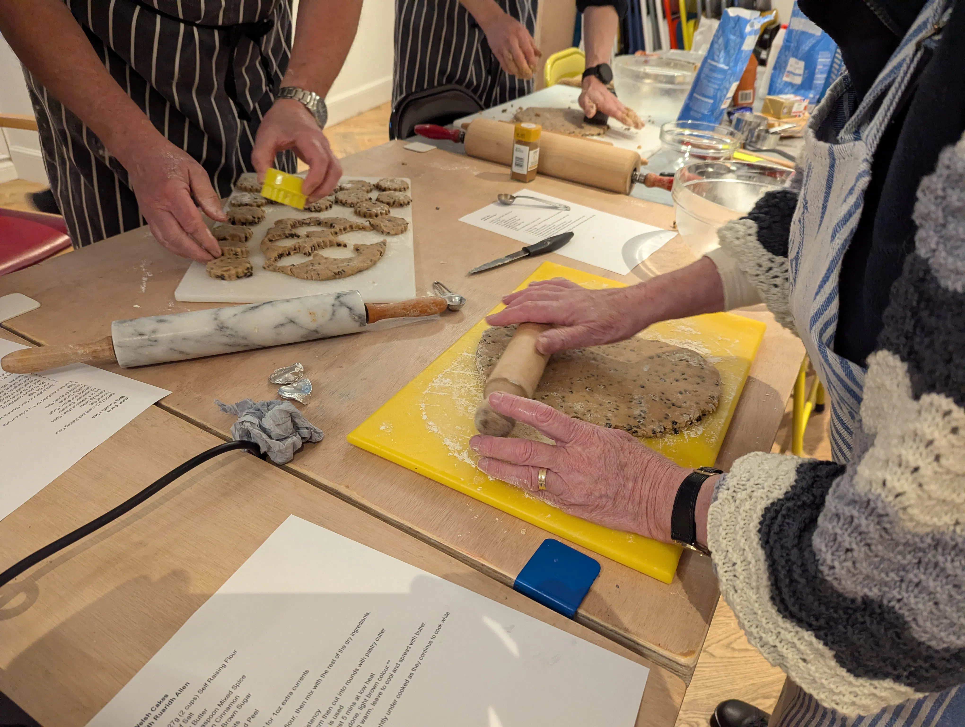 A photo of people working with dough on a wooden table. One person is rolling it out and another cutting circles out. 