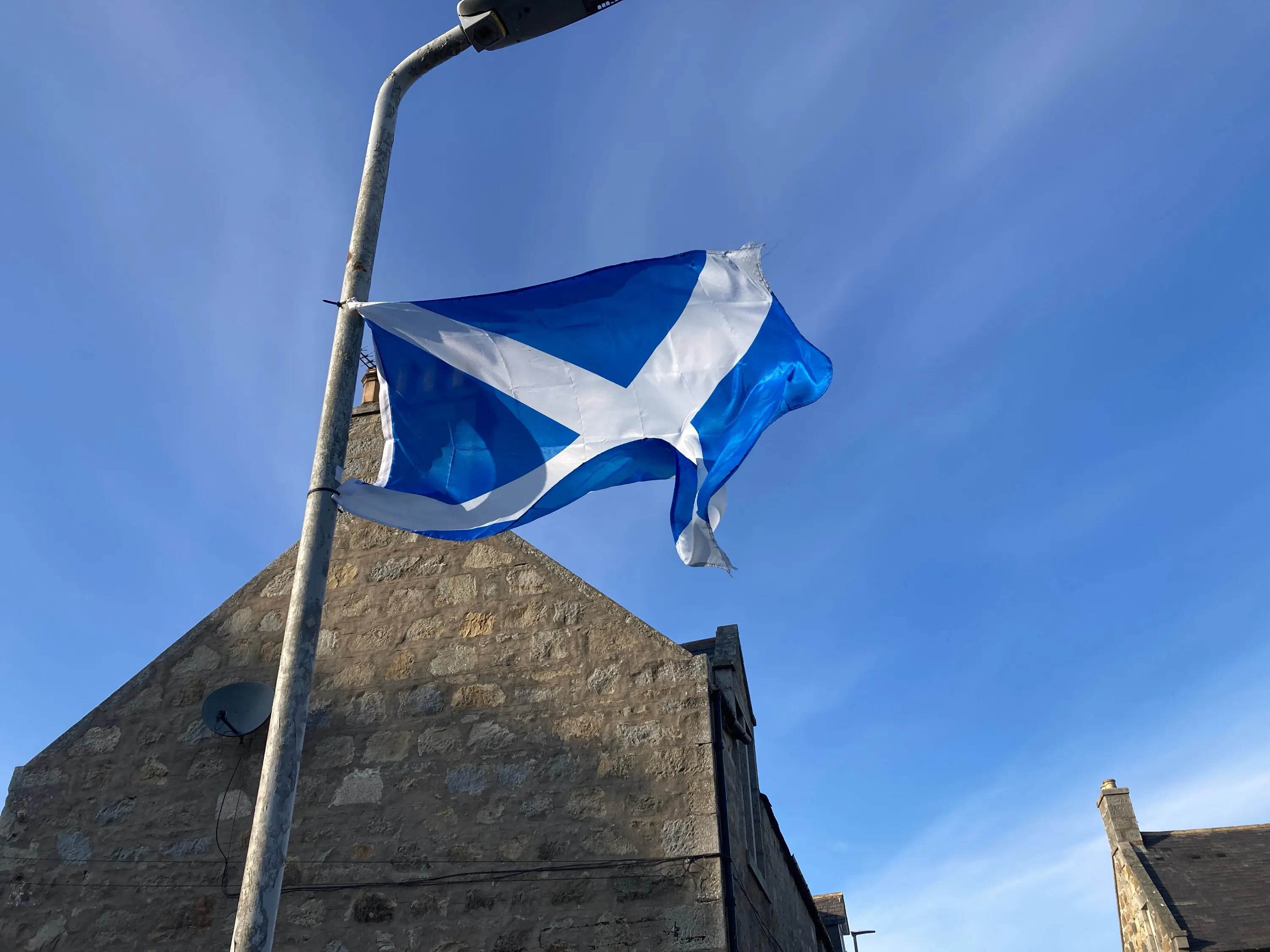 A photo showing the saltire flag, blue with a white cross, blowing in the wind against a blue sky. It is attached to a lamppost and there is an old stone building in the background.