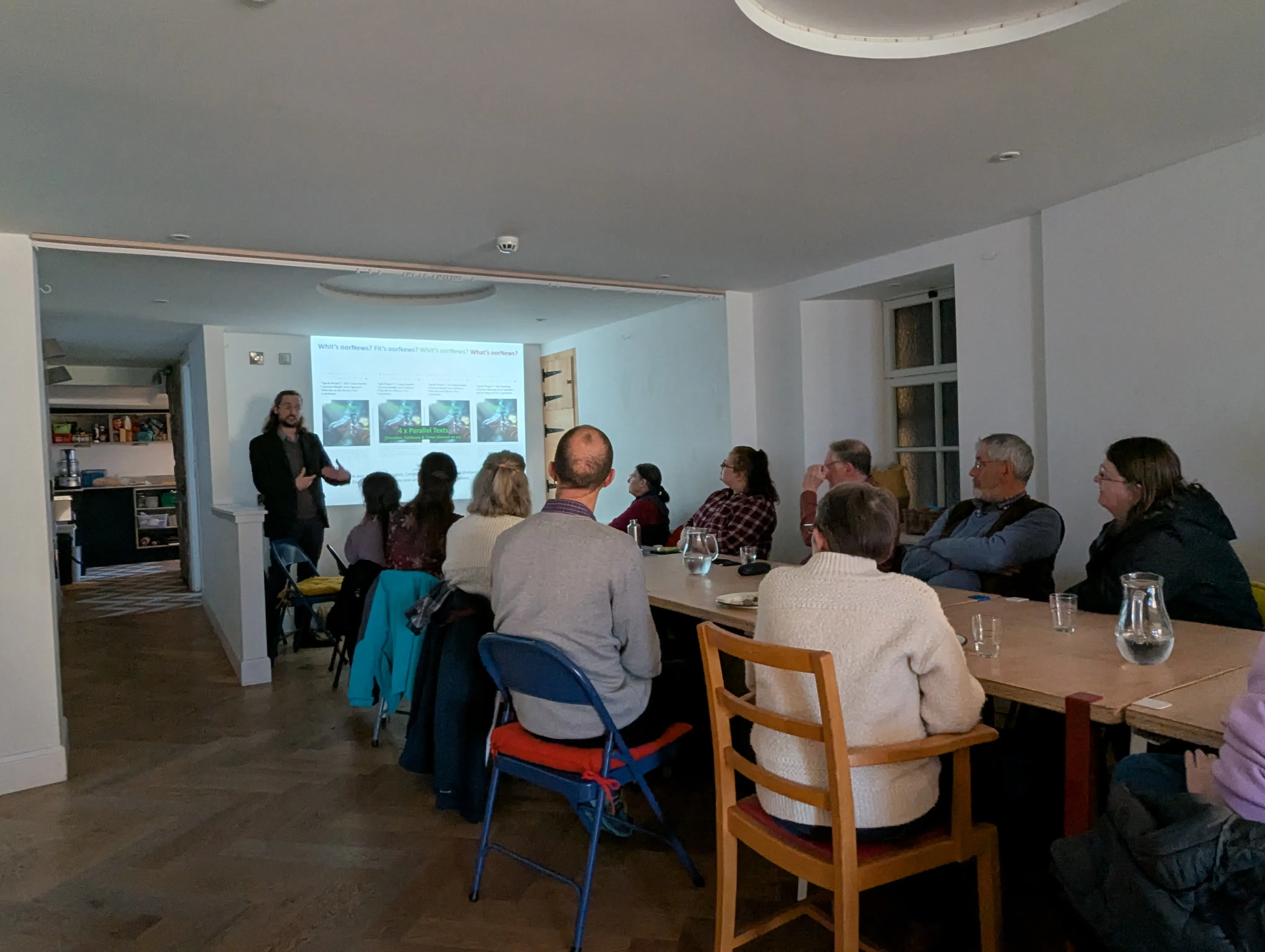 A photo of a person with long brown hair and a beard, wearing a brown suit, presenting to people sat at a long wooden table.