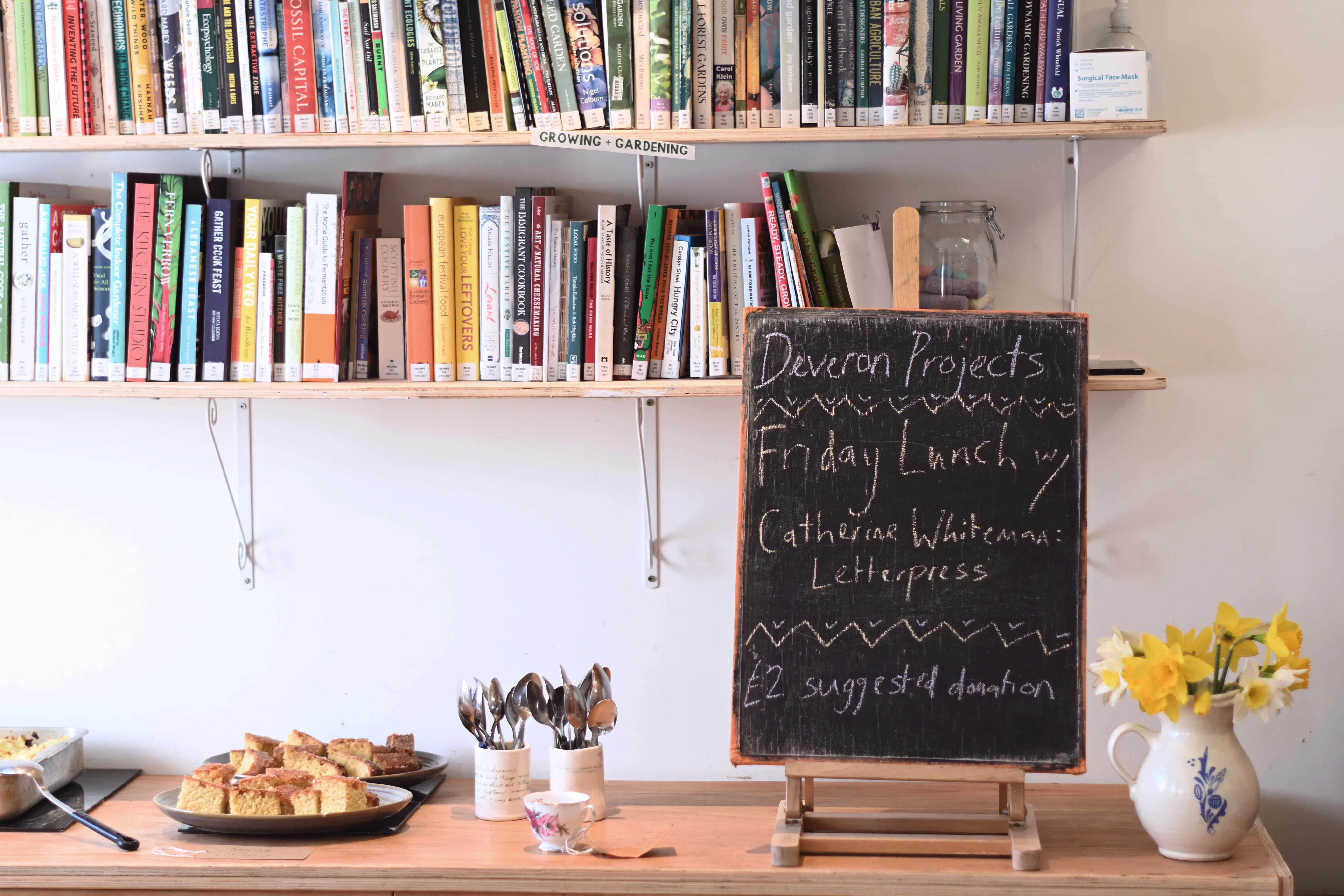 A photo of a small blackboard on an easel with a vase of daffodils and a plate of cake. The blackboard has chalk writing that says Deveron Projects Friday Lunch. 