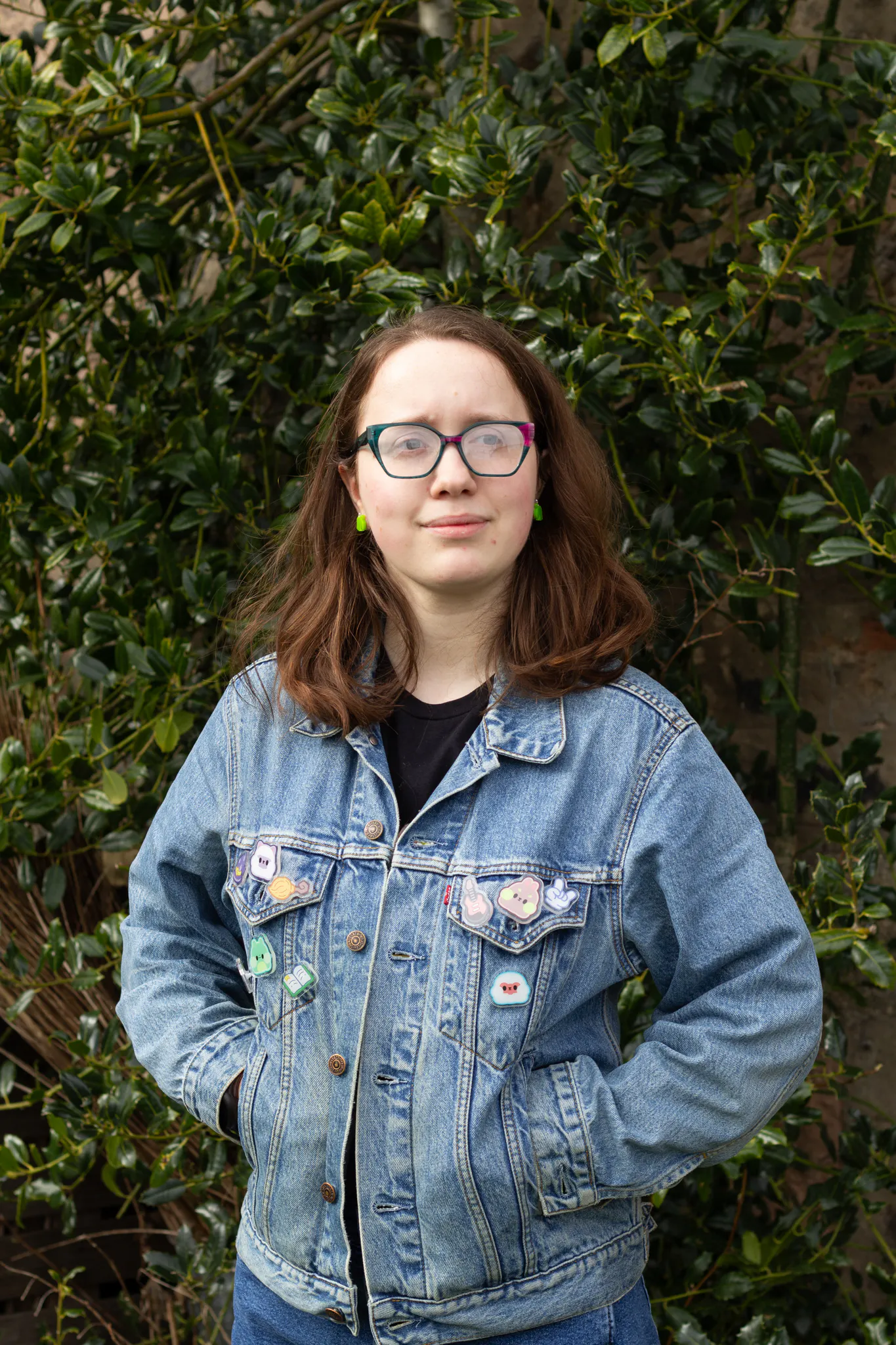Luci has light skin and shoulder length straight dark hair. They are wearing dark glasses and a denim jacket with badges on the pockets.