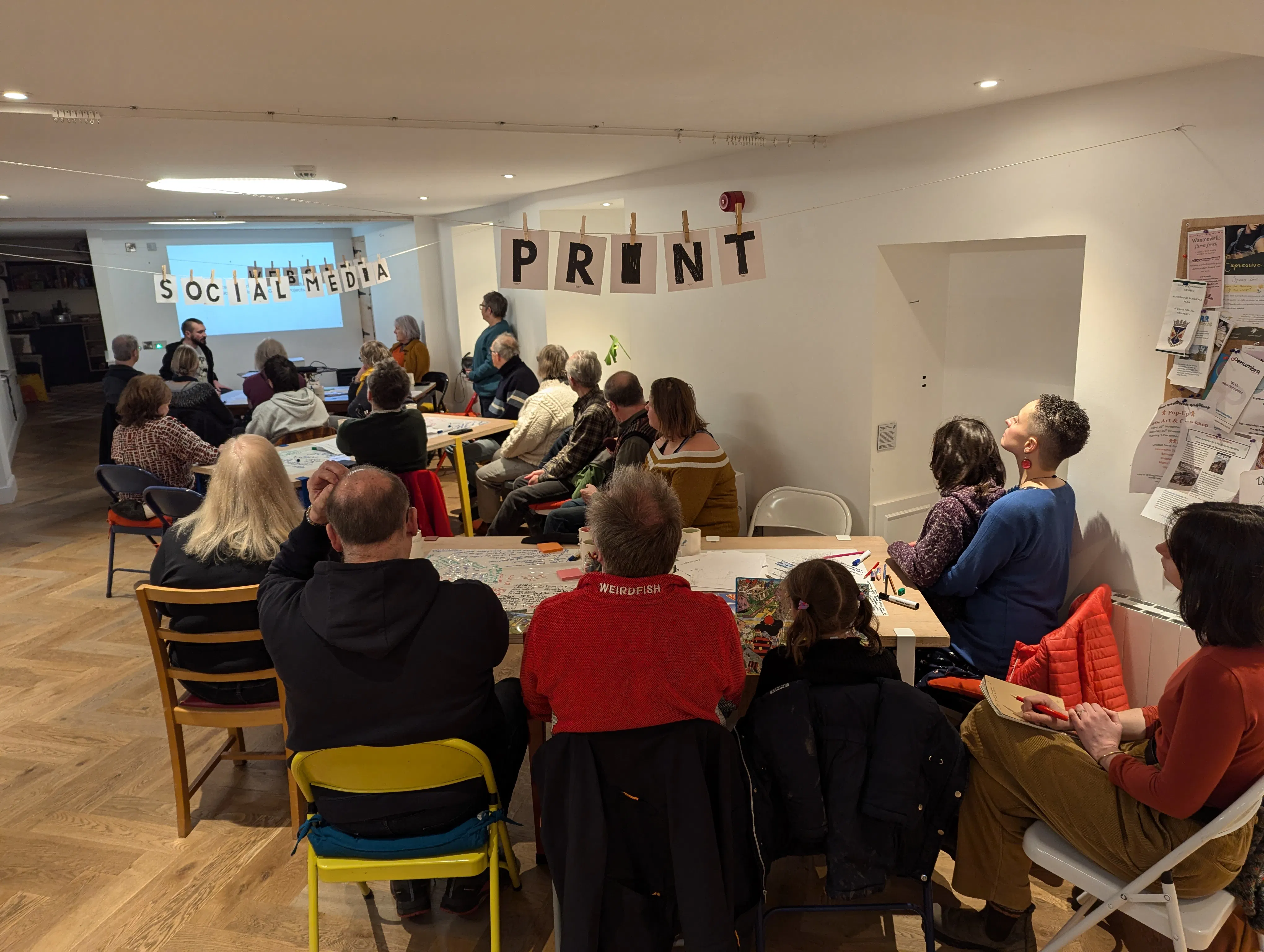 A photo showing around 25 people watching a presentation by a person with dark hair and a beard. There are letters strung across the tables, the front one reading 'print'