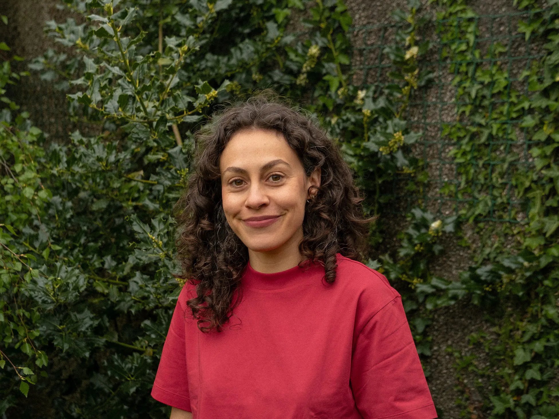 Ane, a white woman with dark brown, curly shoulder-length hair smiles at the camera. She is wearing a red t-shirt and is stood in front of a leafy background.