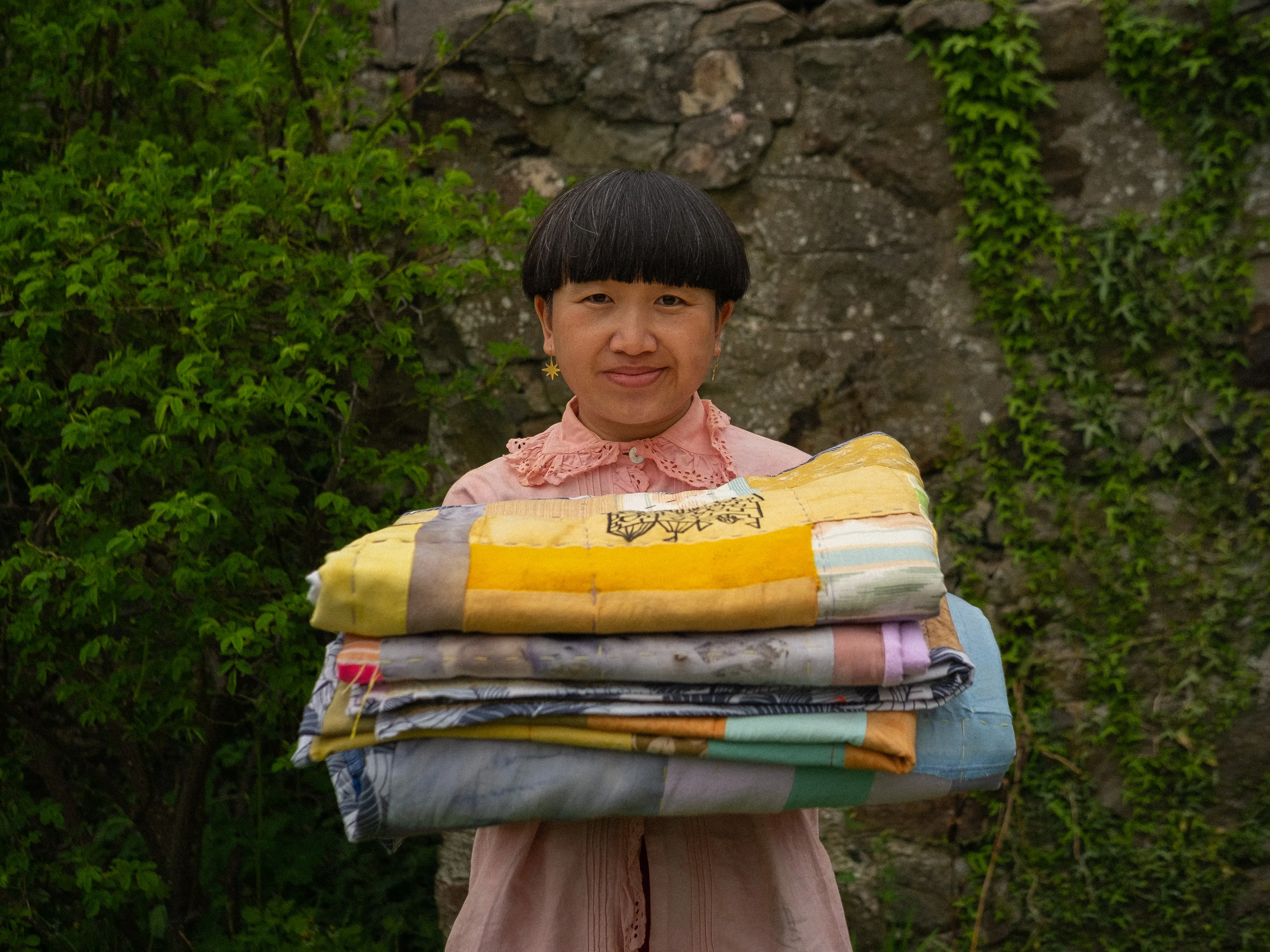 Keng Keng Tang is a south-east Asian woman, with short dark hair in a sharp bob and light brown skin. She is wearing a pink shirt and earrings, and is holding the community quilt in a neatly folded pile.