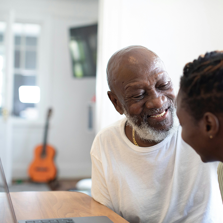 An older man smiling while looking at a laptop computer with a companion indoors.