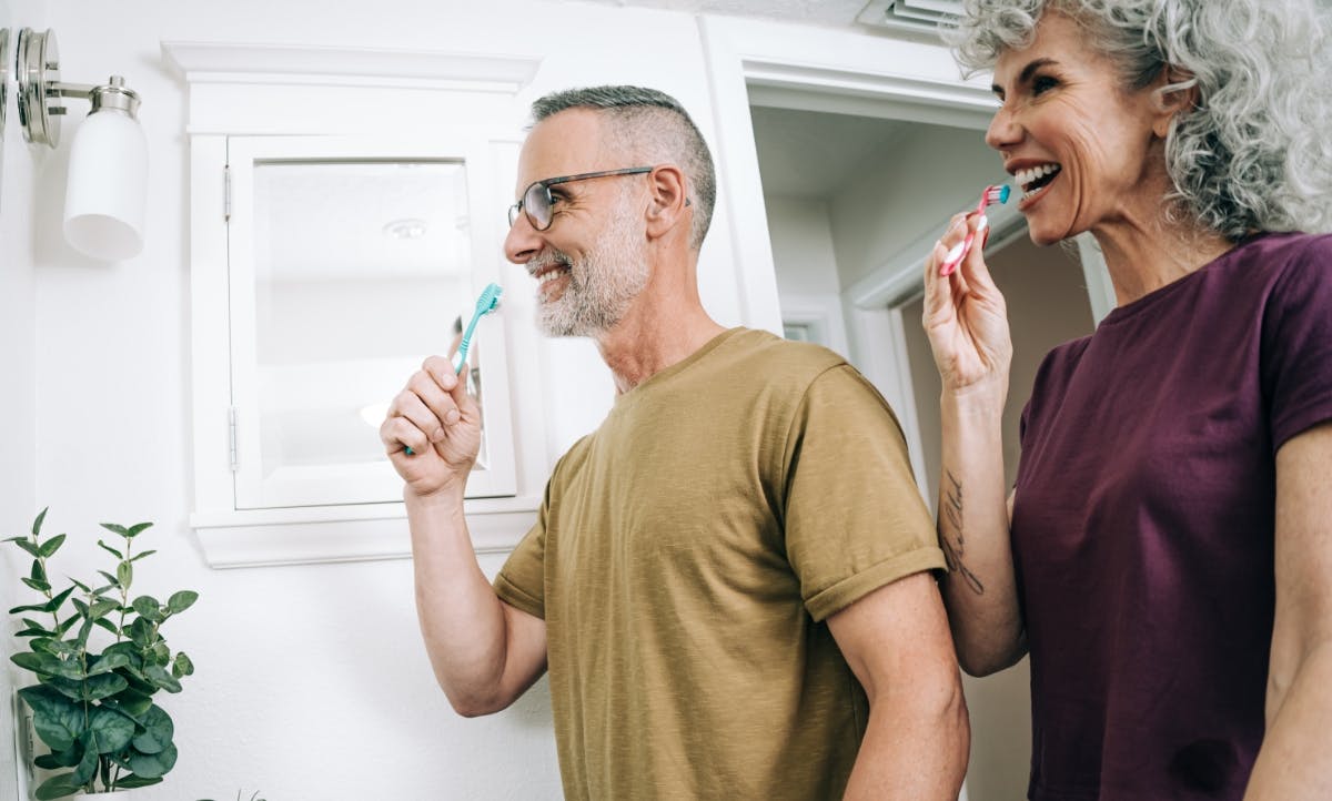 A man and woman brushing their teeth in front of a bathroom mirror.