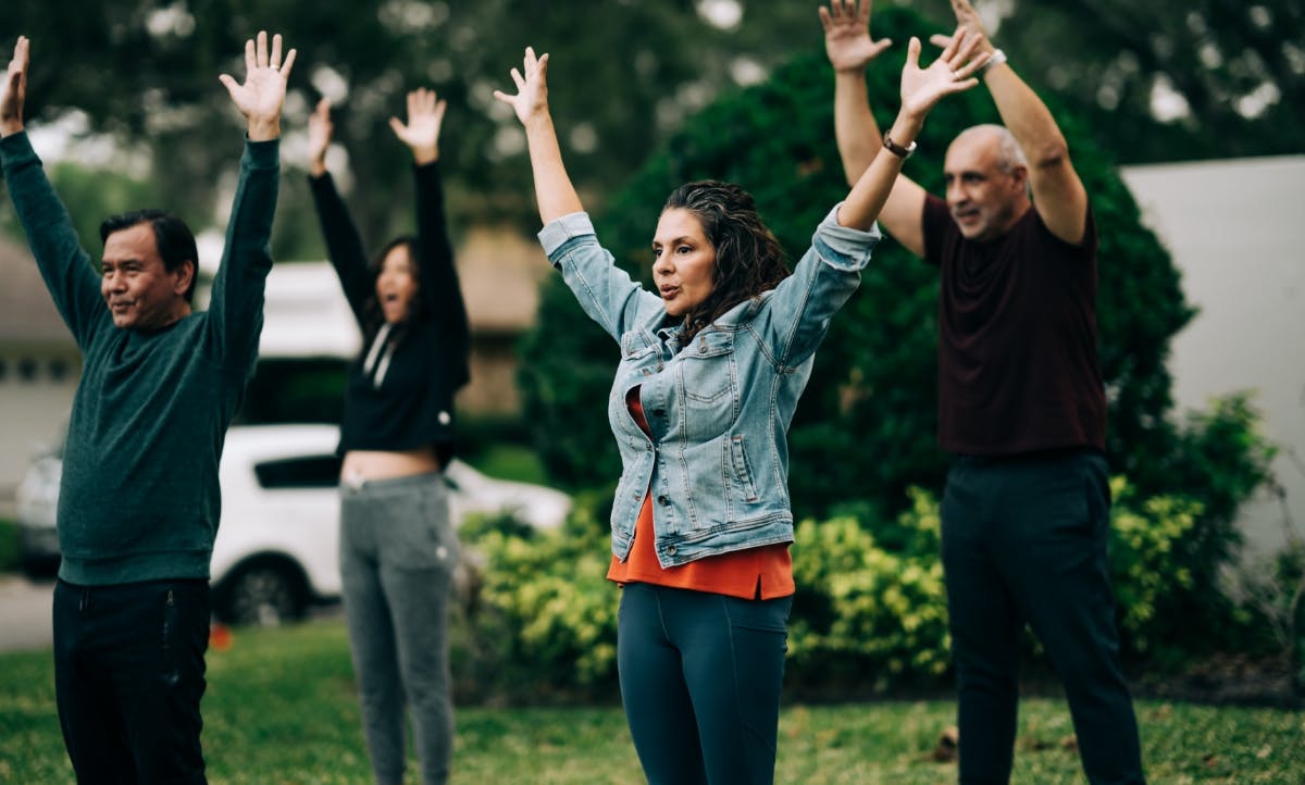 A group of individuals participating in a yoga session outside, standing in a yard with their hands lifted upwards.