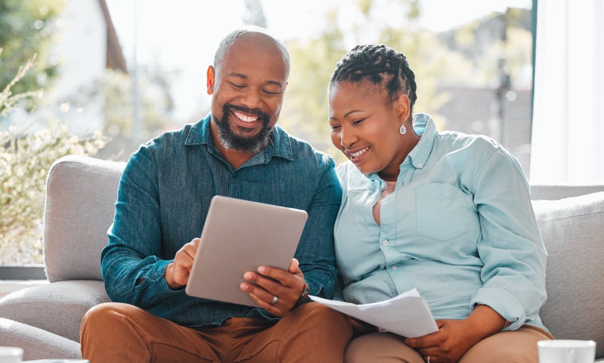 Man and a woman sitting on couch reviewing paperwork and using a tablet.