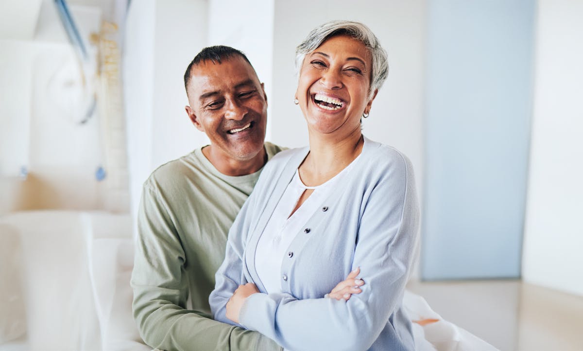 Smiling older couple sitting together indoors, with the woman in front crossing her arms and the man behind her.