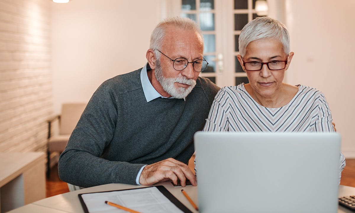 Una pareja de ancianos se sienta uno al lado del otro, comprometidos y sonrientes mientras exploran juntos la pantalla de una computadora portátil.