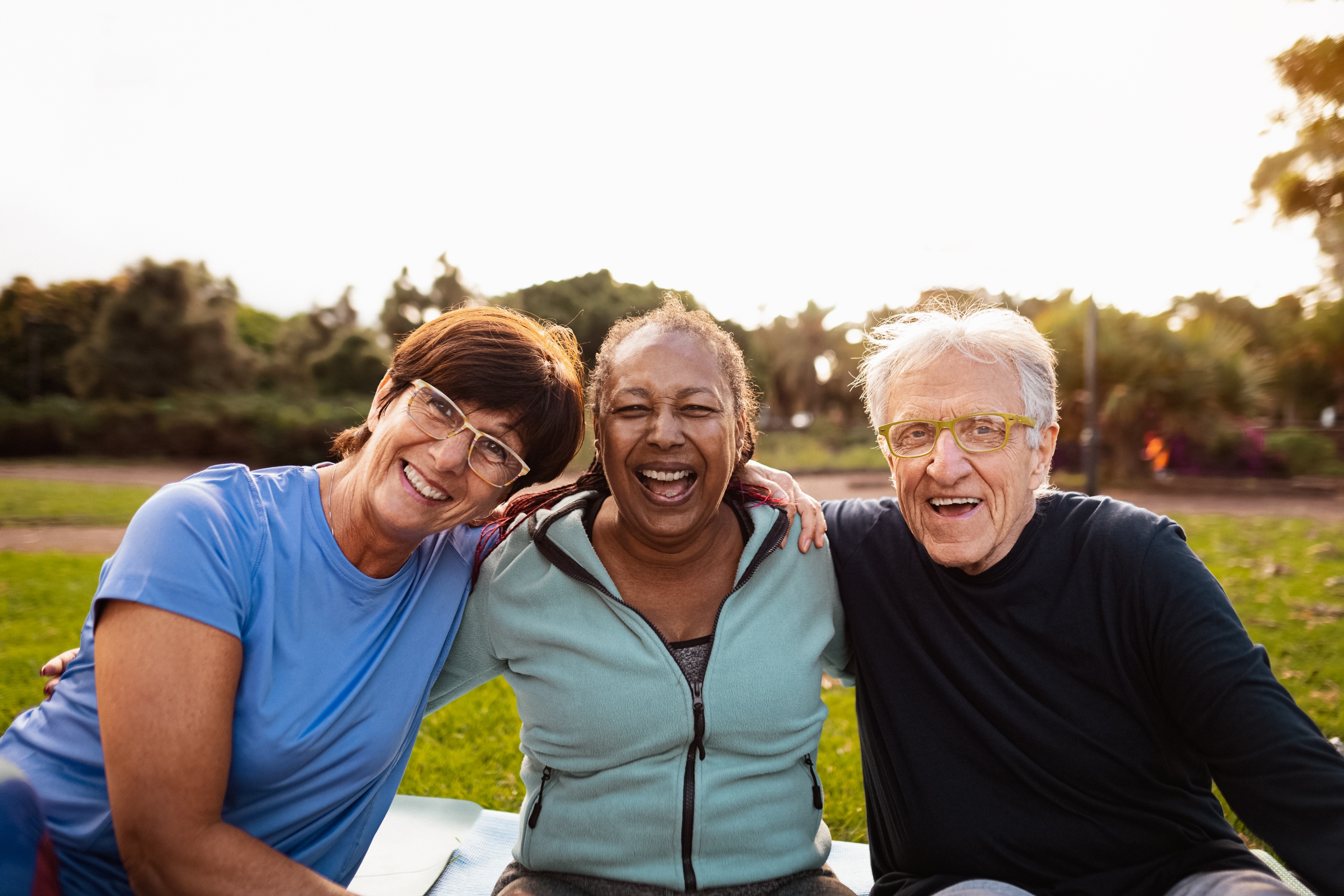 Three older adults relax on a blanket in the park, enjoying the sunny day and each other's company.