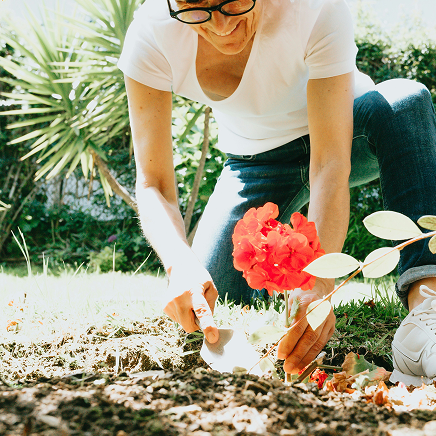 A person kneeling in a garden using a small trowel to plant bright red flowers in the soil.