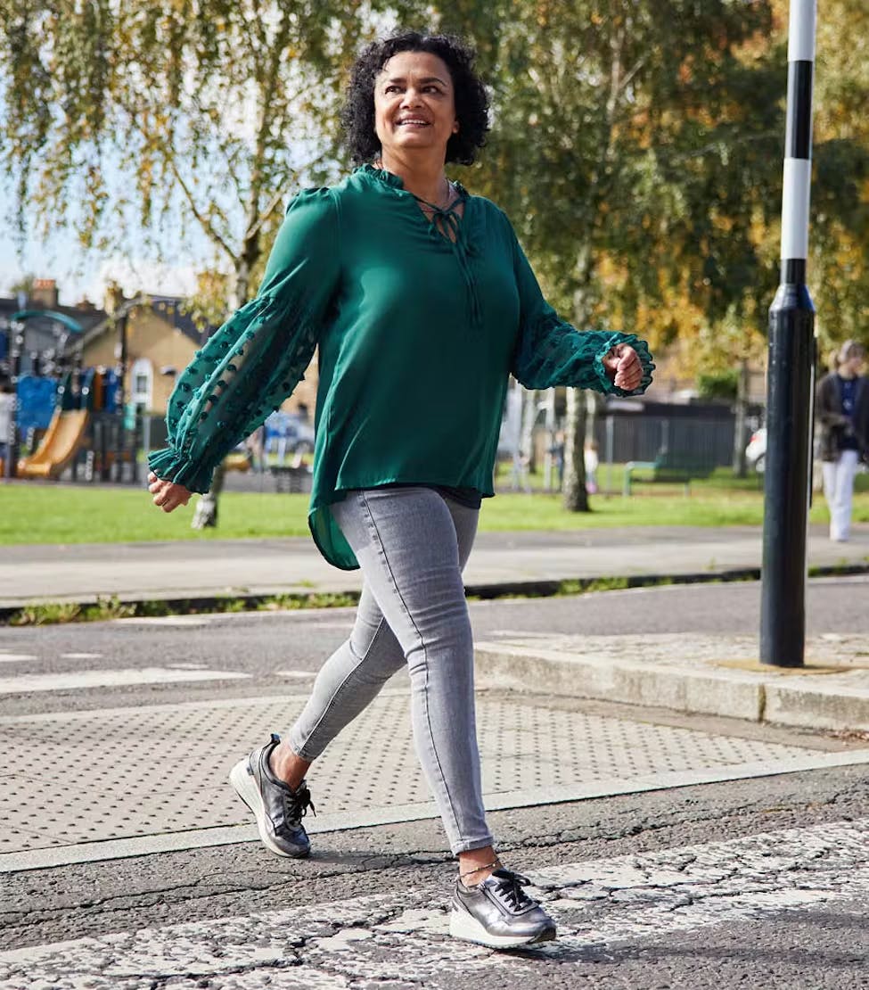 A person walks confidently across a street in a green blouse and grey leggings on a sunny day.