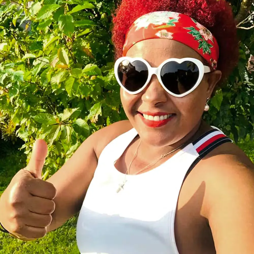 A person outdoors gives a thumbs‑up while wearing a white tank top, a red headband, and sunglasses, standing in bright sunlight near leafy greenery.