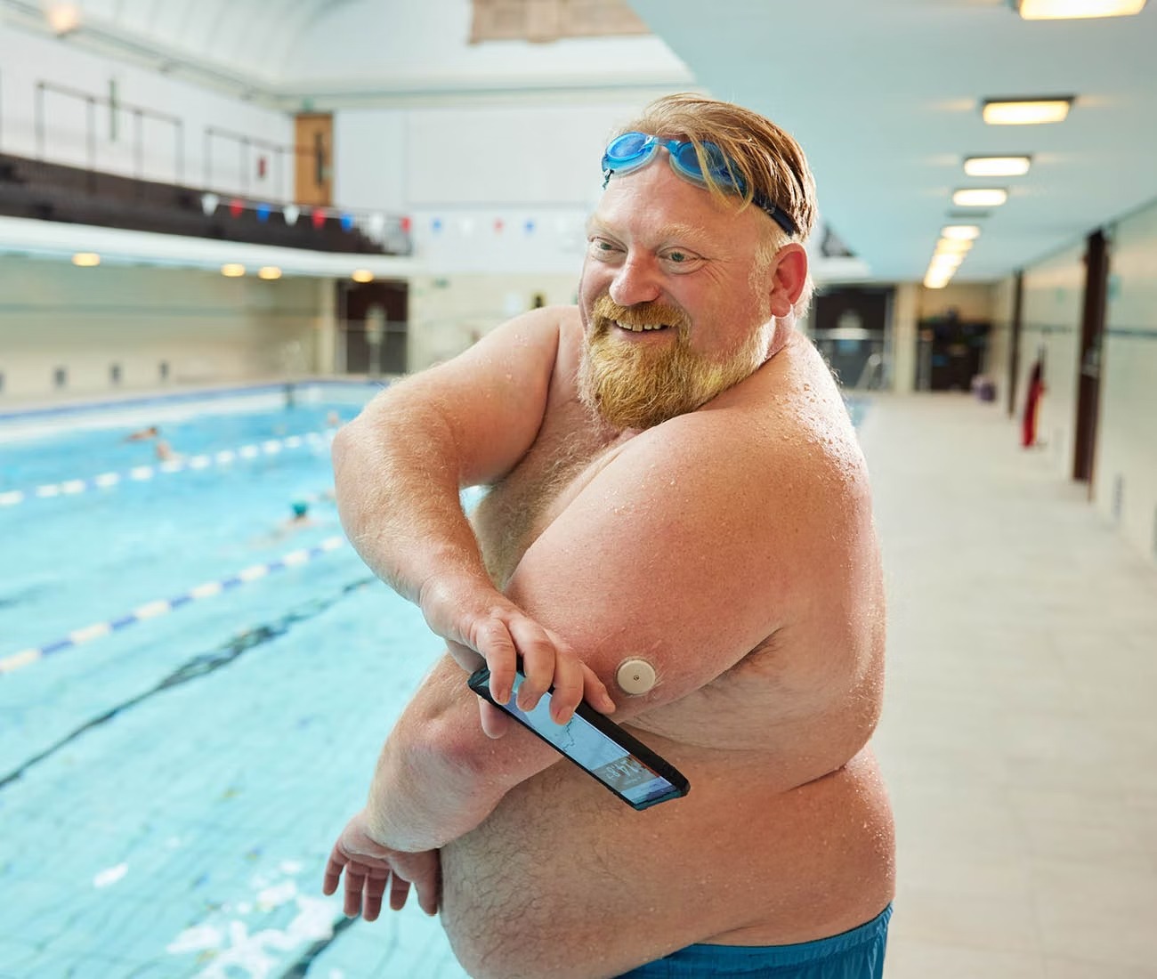 Person with swim goggles and a medical device stands by an indoor pool, holding a smartphone.