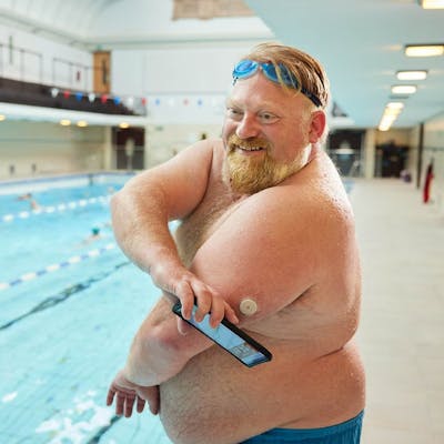 Person with swim goggles and a medical device stands by an indoor pool, holding a smartphone.
