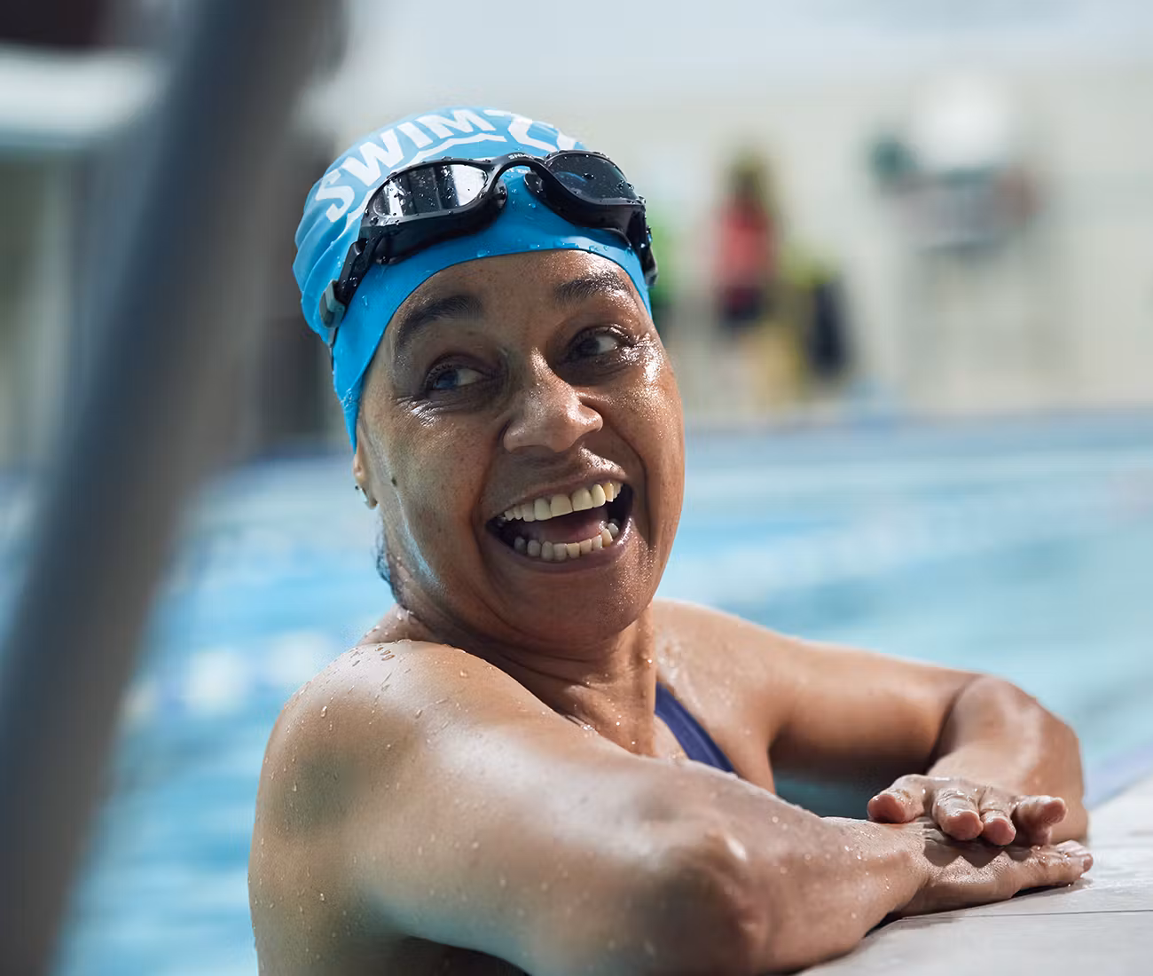 Woman with a swimming hat on and goggles smiling at the edge of a pool