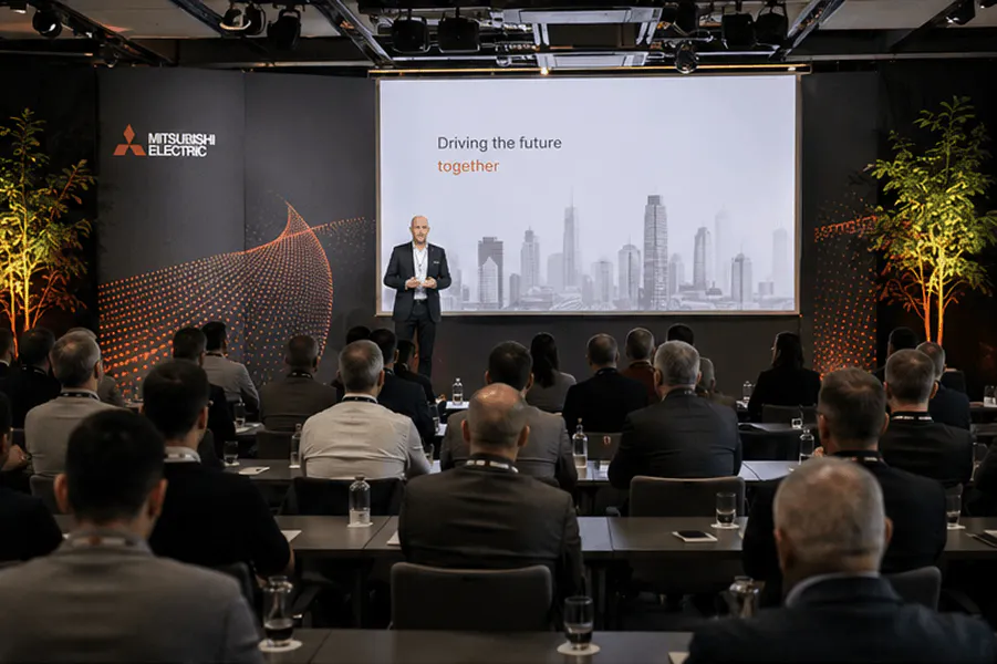 A speaker presents to a room of attendees at a professional conference, with a projection screen in the background.