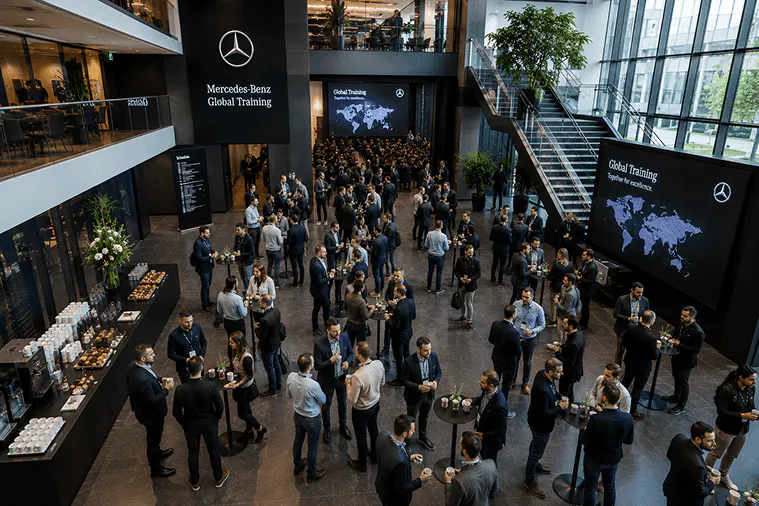 In einem lichtdurchfluteten Atrium mit Empore und Treppe versammeln sich die Teilnehmenden vor Bildschirmen mit dem Veranstaltungsprogramm.