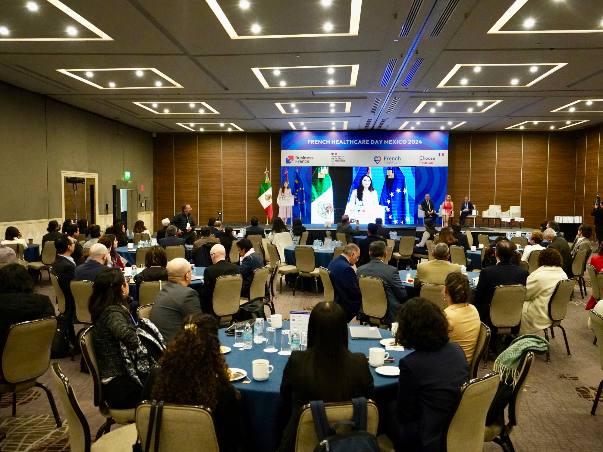 
Vue d’ensemble d’une salle de conférence professionnelle lors du French Healthcare Day Mexico 2024, avec des participants assis à des tables rondes, une scène avec intervenants et écrans, et les drapeaux du Mexique, de la France et de l’Union européenne en arrière-plan.