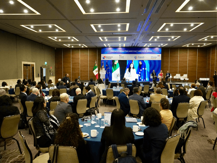 Vue d’ensemble d’une salle de conférence professionnelle lors du French Healthcare Day Mexico 2024, avec des participants assis à des tables rondes, une scène avec intervenants et écrans, et les drapeaux du Mexique, de la France et de l’Union européenne en arrière-plan.